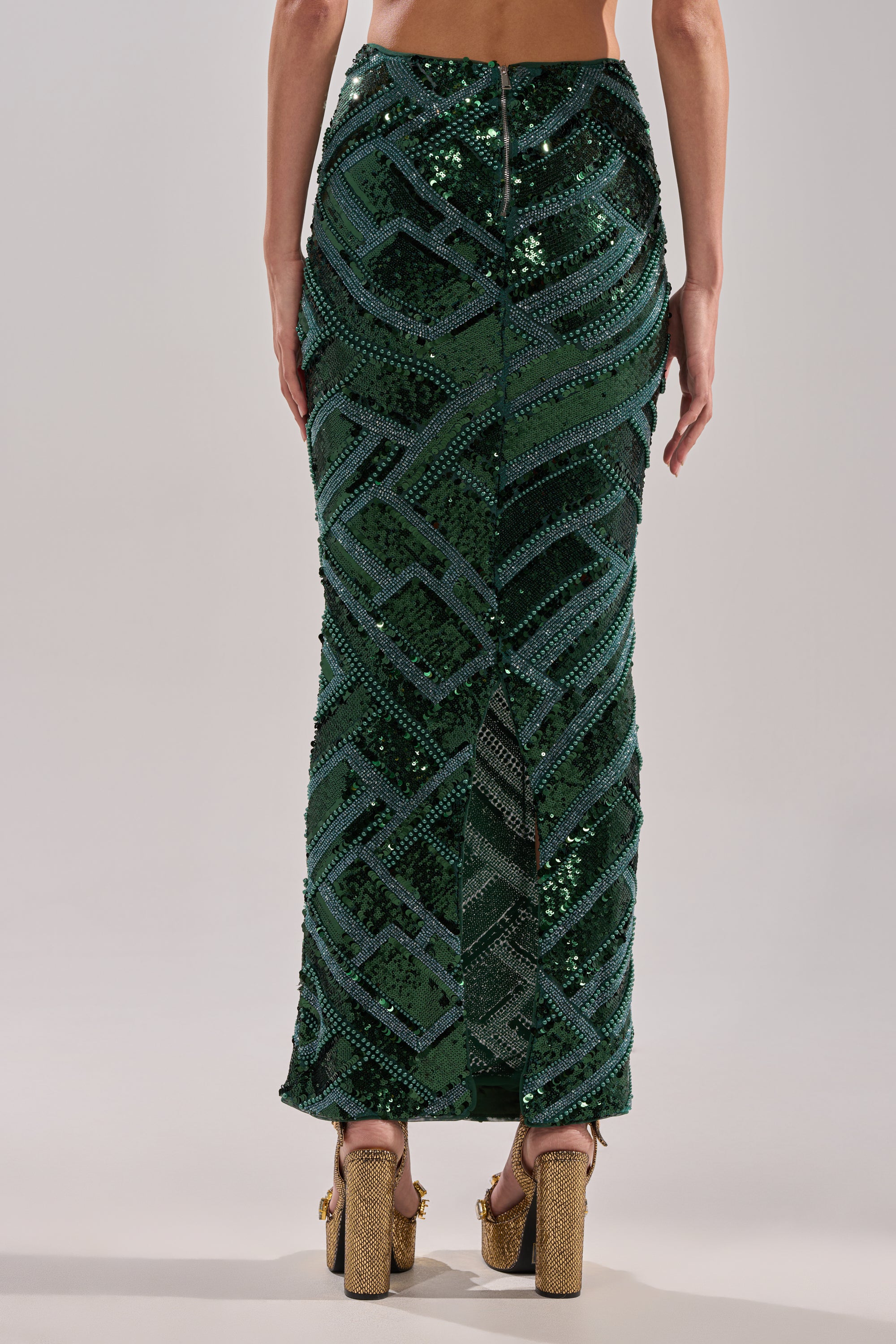 A person wearing the HOTTEST IN THE CLUB SEQUIN MAXI SKIRT, a fitted floor-length maxi with striking geometric sequins and a mermaid silhouette, is seen from the back paired with gold high-heeled shoes.