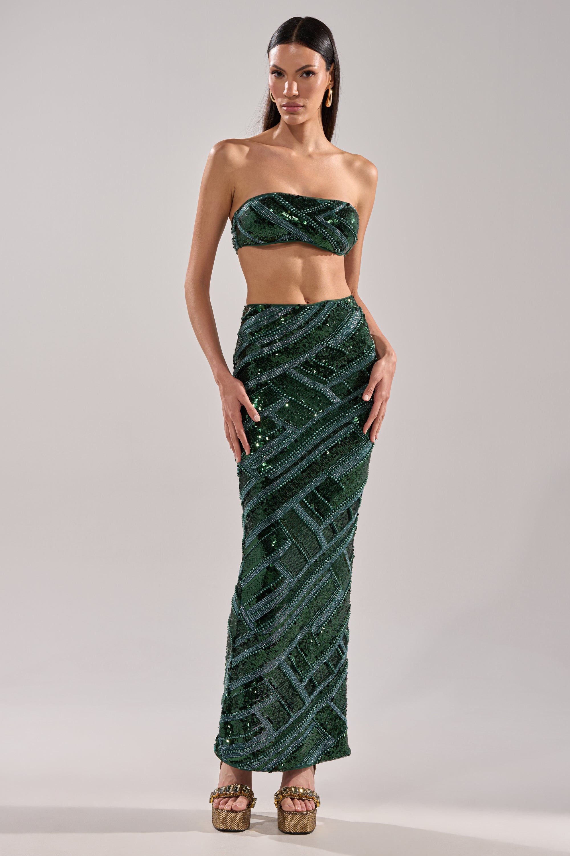 A woman in the HOTTEST IN THE CLUB SEQUIN MAXI SKIRT—a dark green, sequined mermaid silhouette maxi—stands facing forward with long dark hair and platform shoes against a plain light gray background.