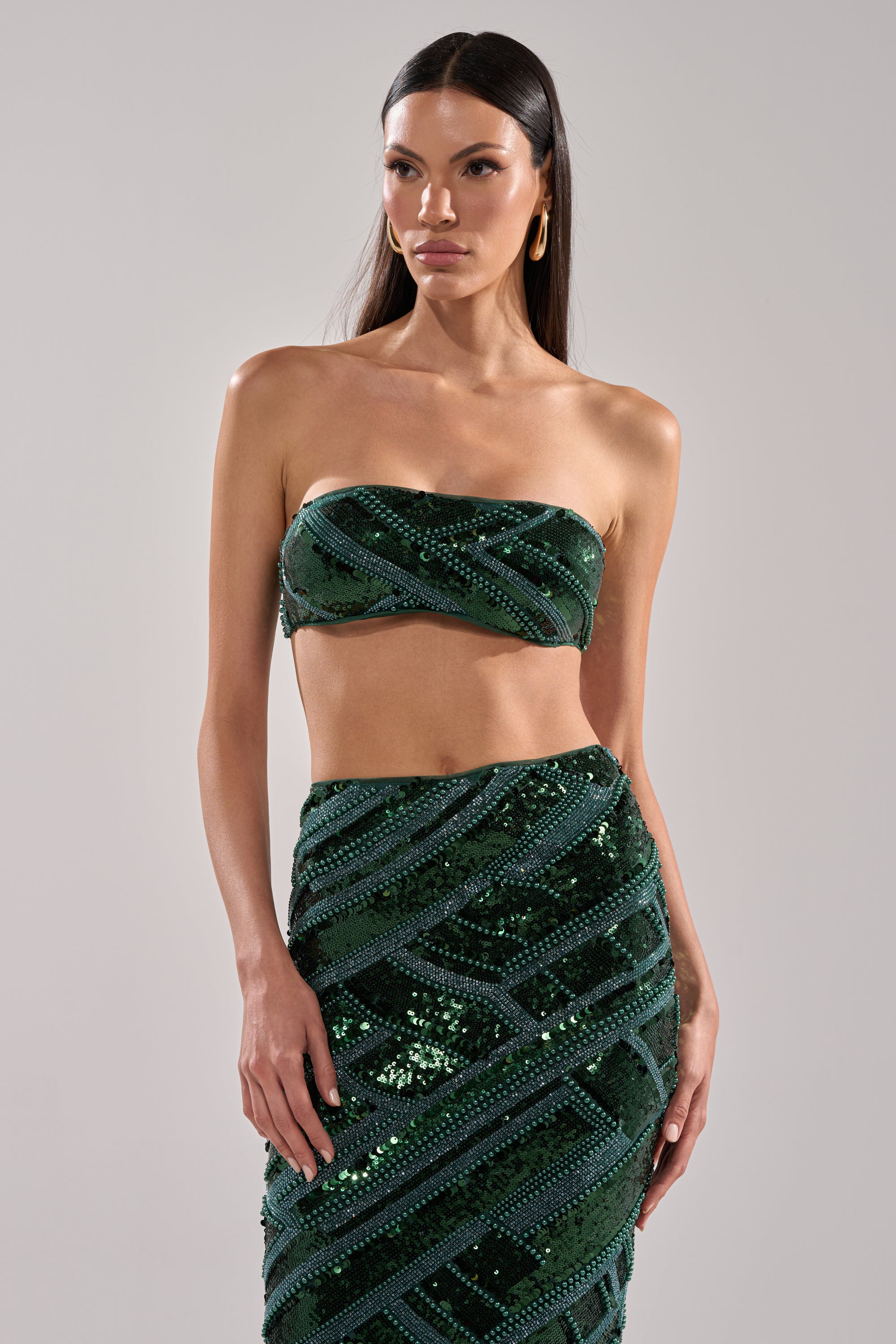 A woman with long dark hair wears the HOTTEST GIRL IN THE CLUB SEQUIN BANDEAU and a matching skirt, posing against a plain light gray background.