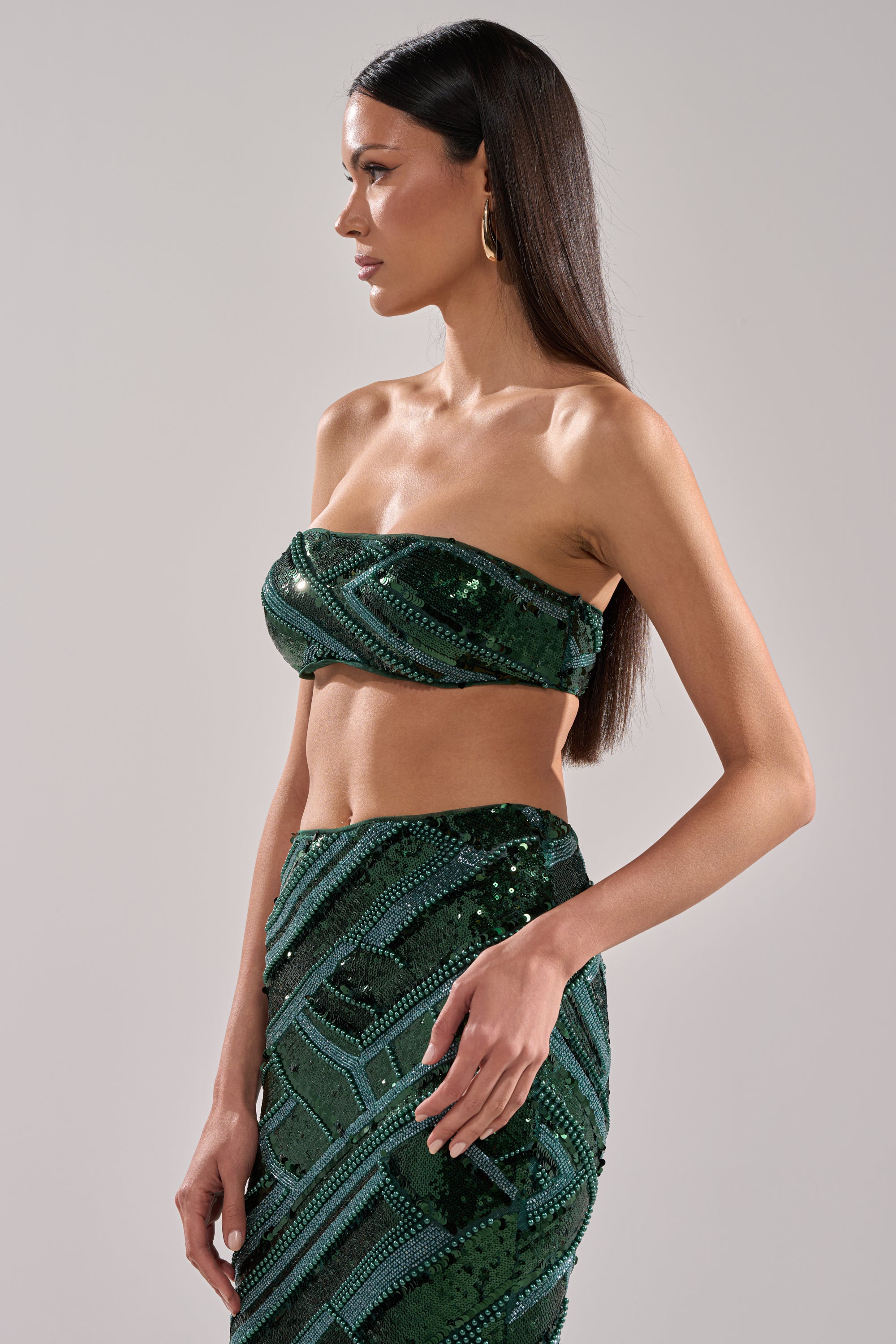 Woman with long straight brown hair wears the HOTTEST GIRL IN THE CLUB SEQUIN BANDEAU and matching high-waisted skirt, shown in profile against a plain light background. She has gold hoop earrings and is looking to the side.