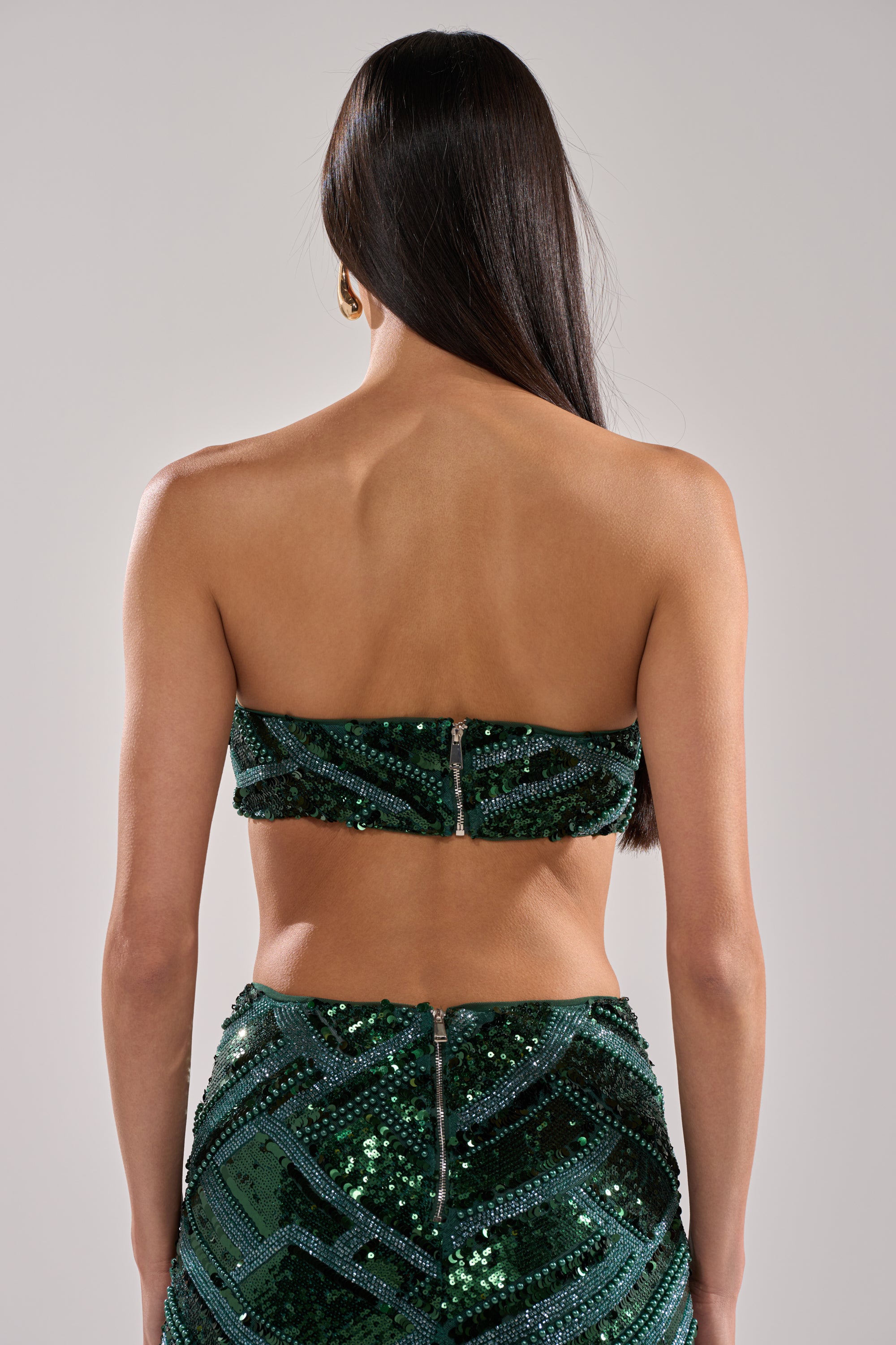 A woman with long straight hair stands facing away, wearing the HOTTEST GIRL IN THE CLUB SEQUIN BANDEAU in dark green and a matching high-waisted skirt with zipper details, set against a plain light background.