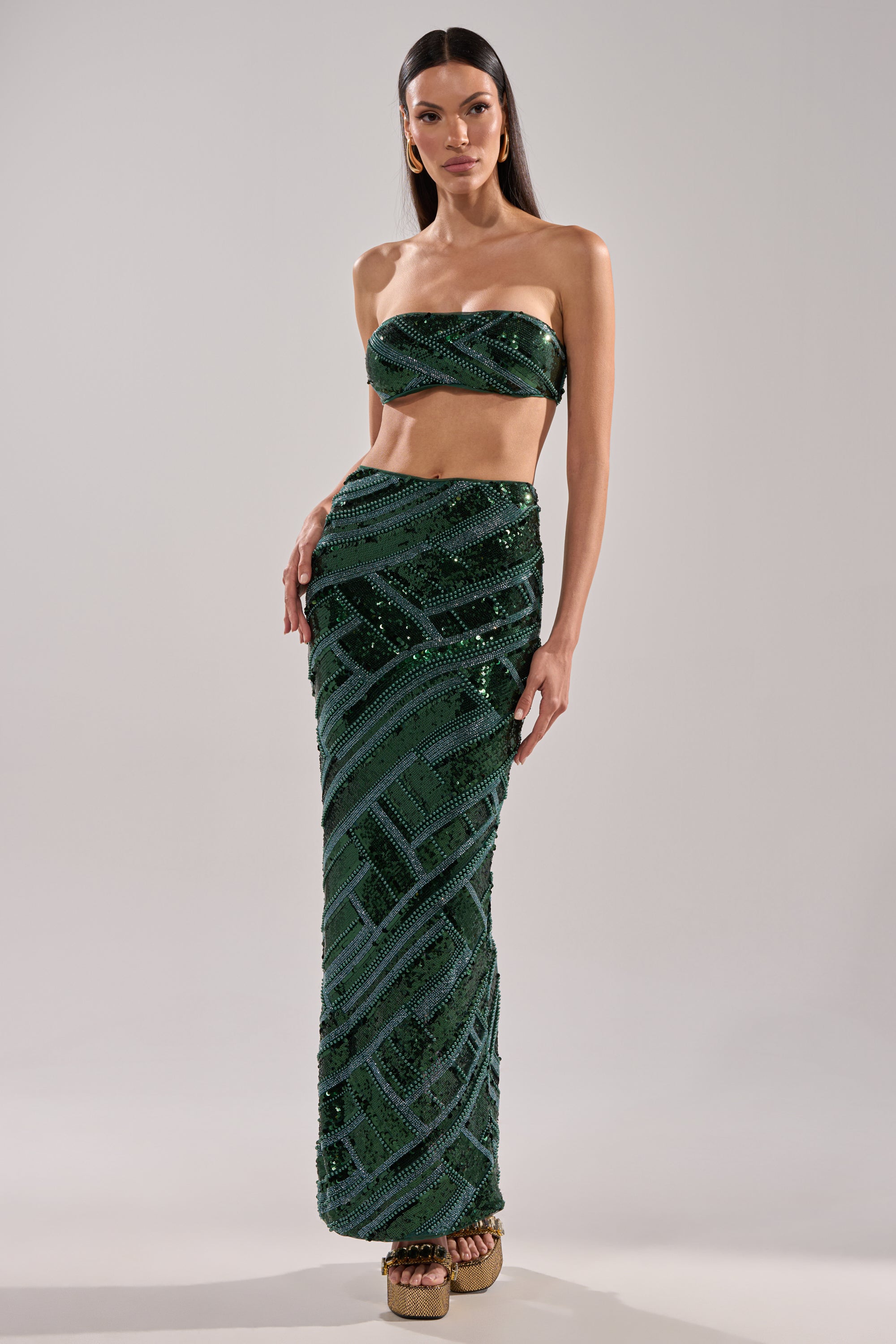 A woman in the HOTTEST GIRL IN THE CLUB SEQUIN BANDEAU and matching geometric high-waisted, floor-length skirt poses against a plain background, her long straight hair styled sleekly, wearing platform sandals.