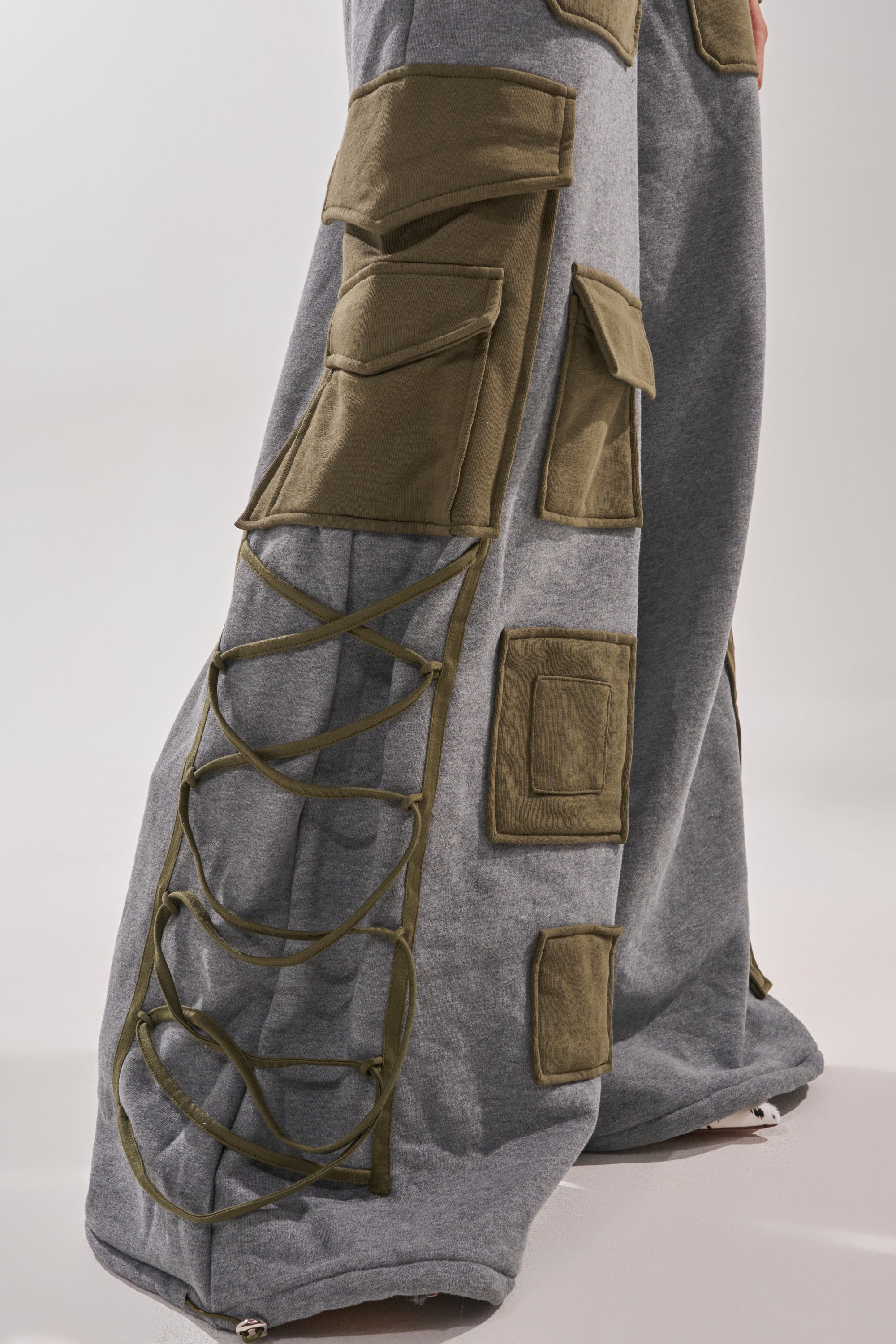 A close-up of the CRAZY IN LOVE TROUSER shows gray high-waisted cargo pants with olive green pockets and crisscross laces on the sides, merging utilitarian style with a unique, textured, and layered look.