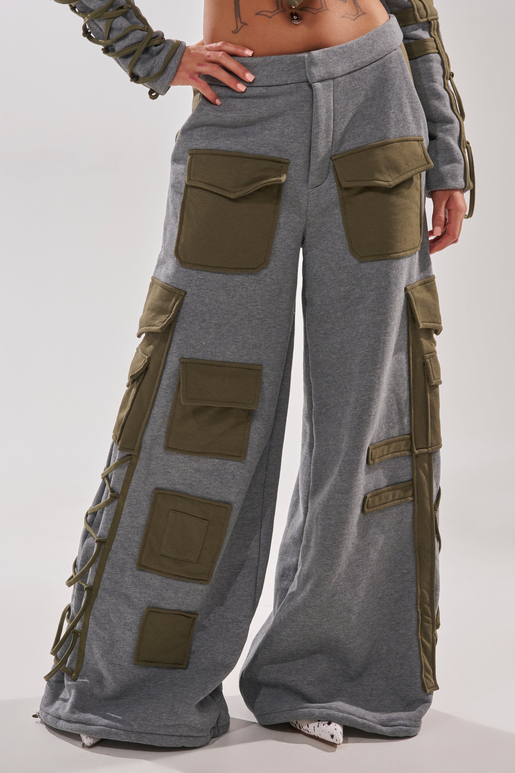 A person wears the CRAZY IN LOVE TROUSER—gray, high-waisted, wide-leg pants with cargo pockets and lace-up side detailing—paired with a cropped top that shows their midriff.
