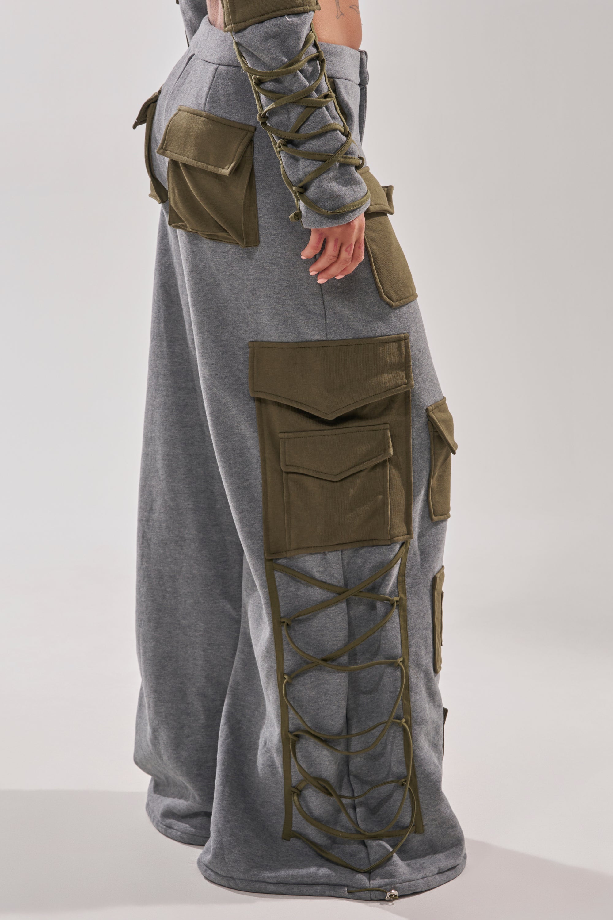 A person wears the CRAZY IN LOVE TROUSER, a high-waisted gray wide-leg pant with large olive cargo pockets and crisscross lace-up sides, paired with a matching sleeve featuring similar laces and cargo pockets.