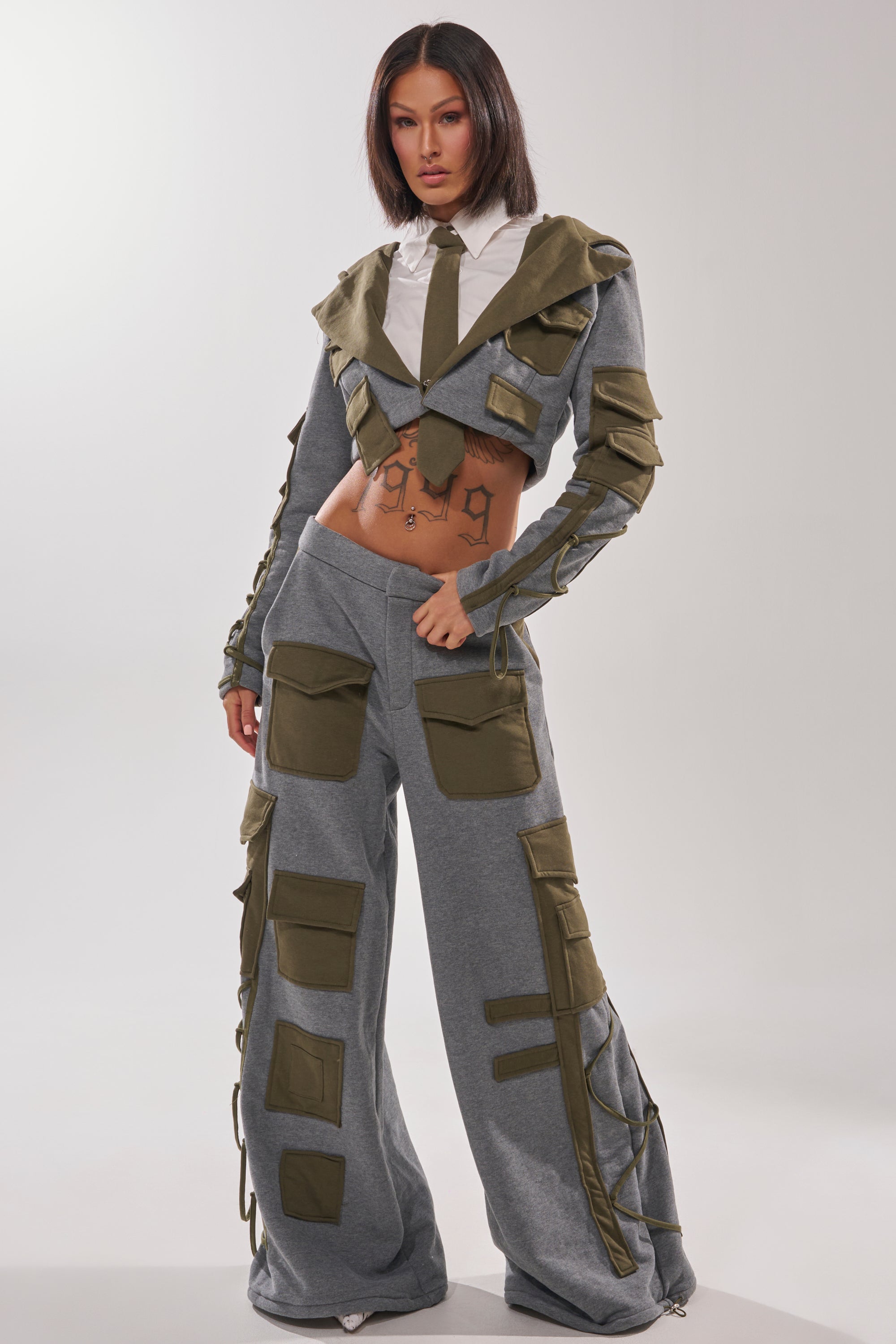 A woman models the CRAZY IN LOVE TROUSER—a gray wide-leg pant with olive green pockets—paired with a cropped jacket, white shirt, and tie. Her midriff is exposed, highlighting tattoos on her stomach and hands.