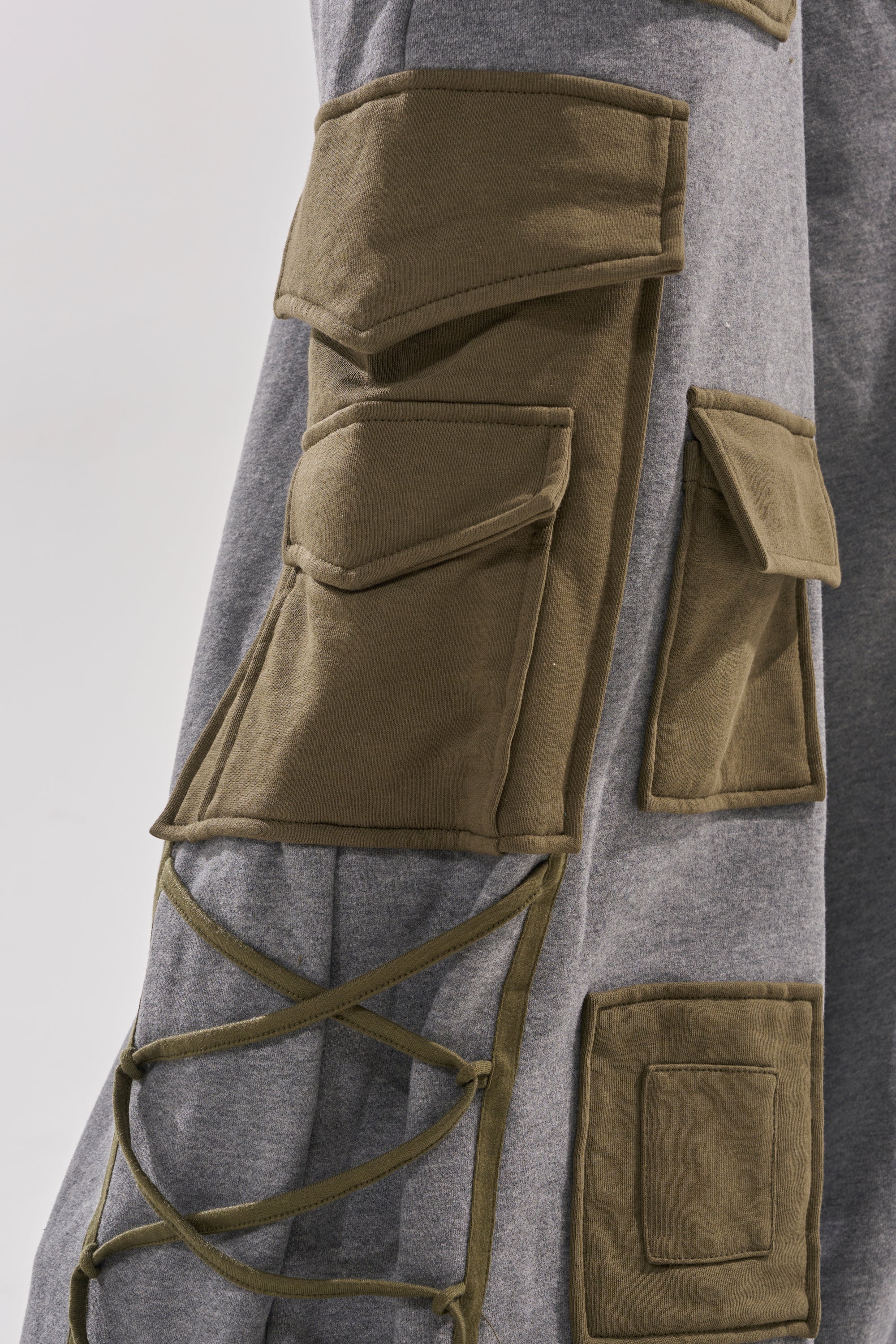 A close-up of the CRAZY IN LOVE TROUSER showcases grey fabric with multiple olive-green pockets and crisscrossed laces, capturing the spirit of utilitarian fashion.