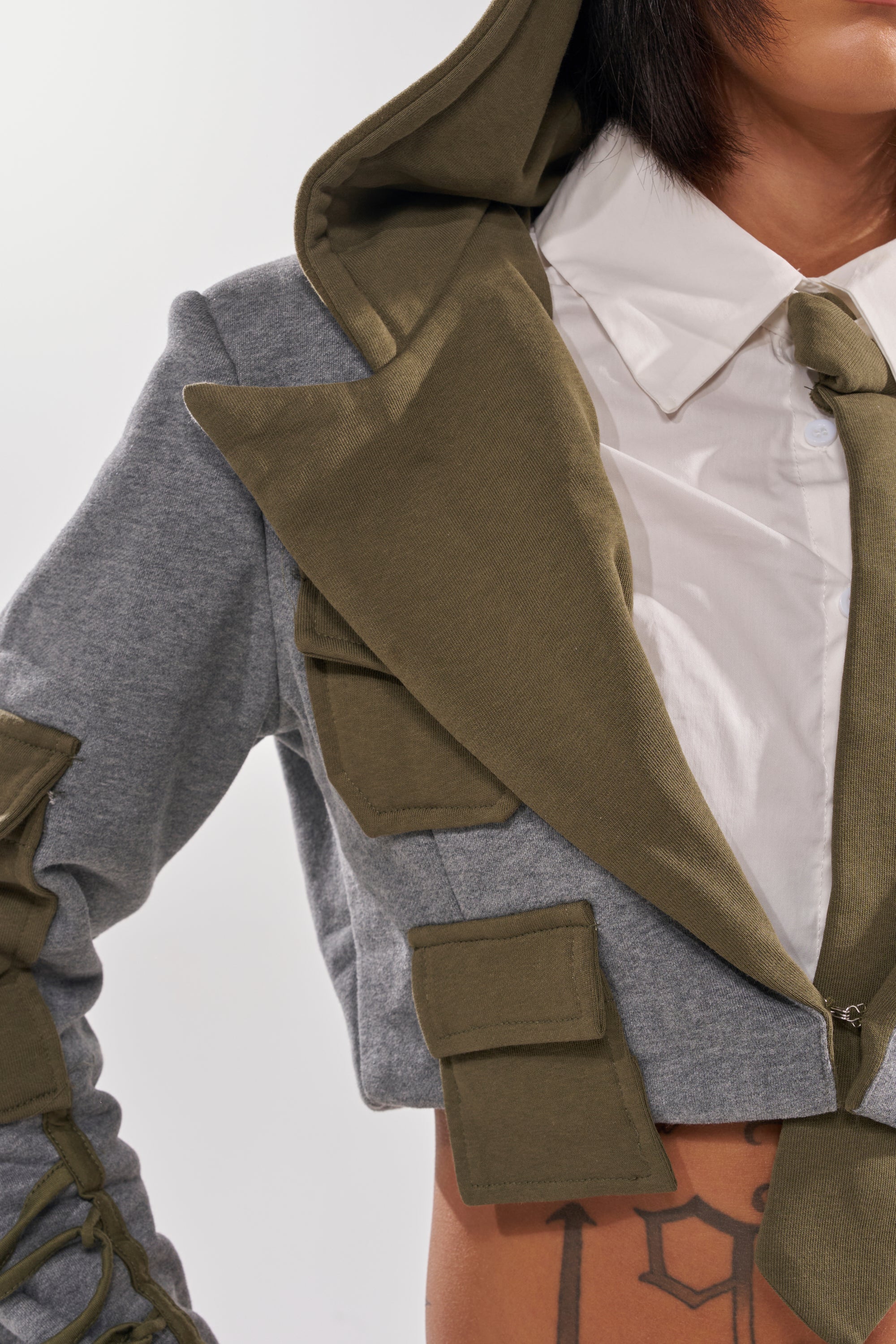 A person wears the CRAZY IN LOVE BLOUSE—a gray and olive green hooded jacket with utility pockets—over a white collared shirt and olive tie. Their torso has visible tattoos, while their face is mostly out of frame.