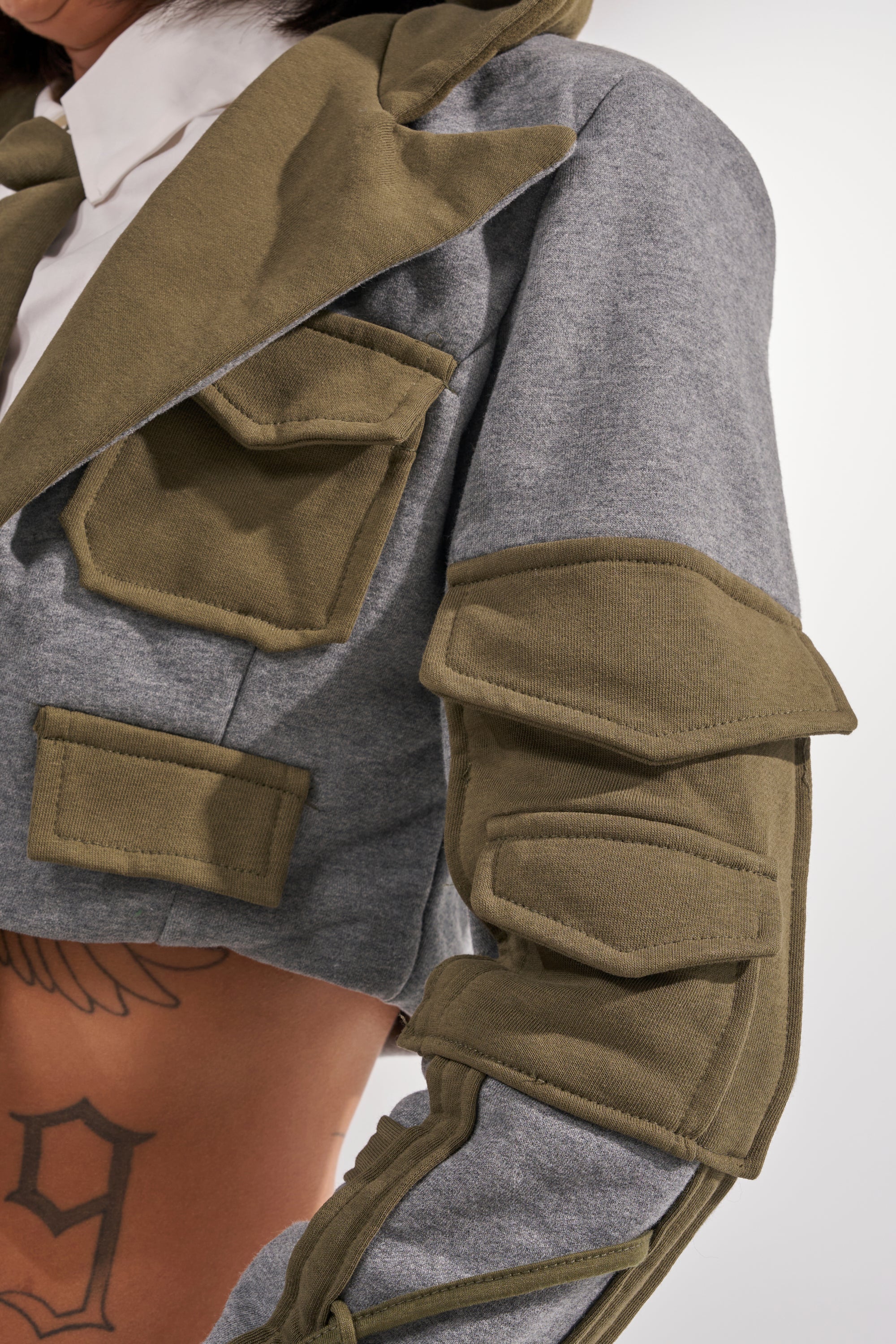 A close-up of a person wearing the CRAZY IN LOVE BLOUSE—a cropped gray jacket with olive-green utility pockets and patches on the front and sleeve. The streetwear top reveals their midriff and a partially visible tattoo.