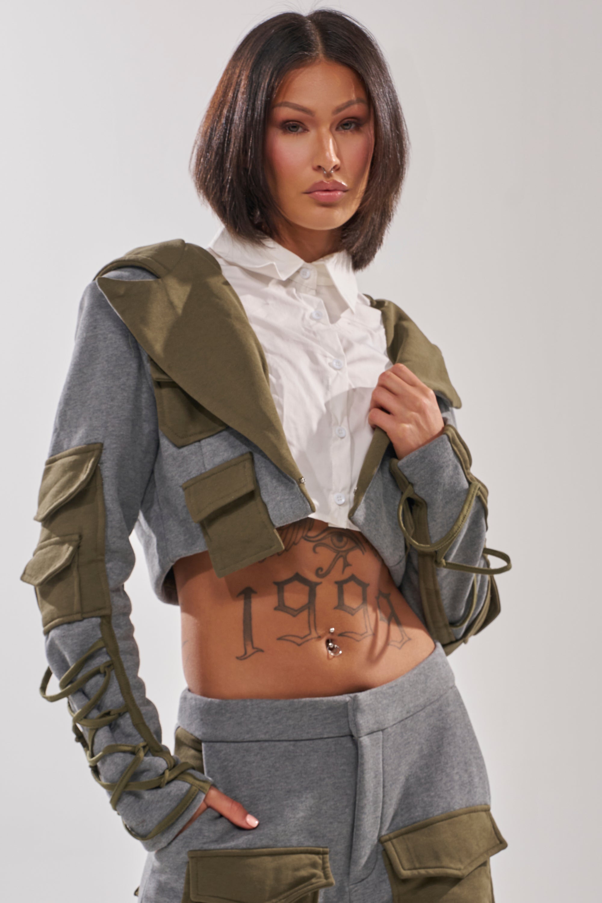 A woman with a bob wears the CRAZY IN LOVE BLOUSE—a cropped, grey and olive green jacket with utility pockets over a white shirt—showing a "1909" stomach tattoo, matching pants, and a confident look.