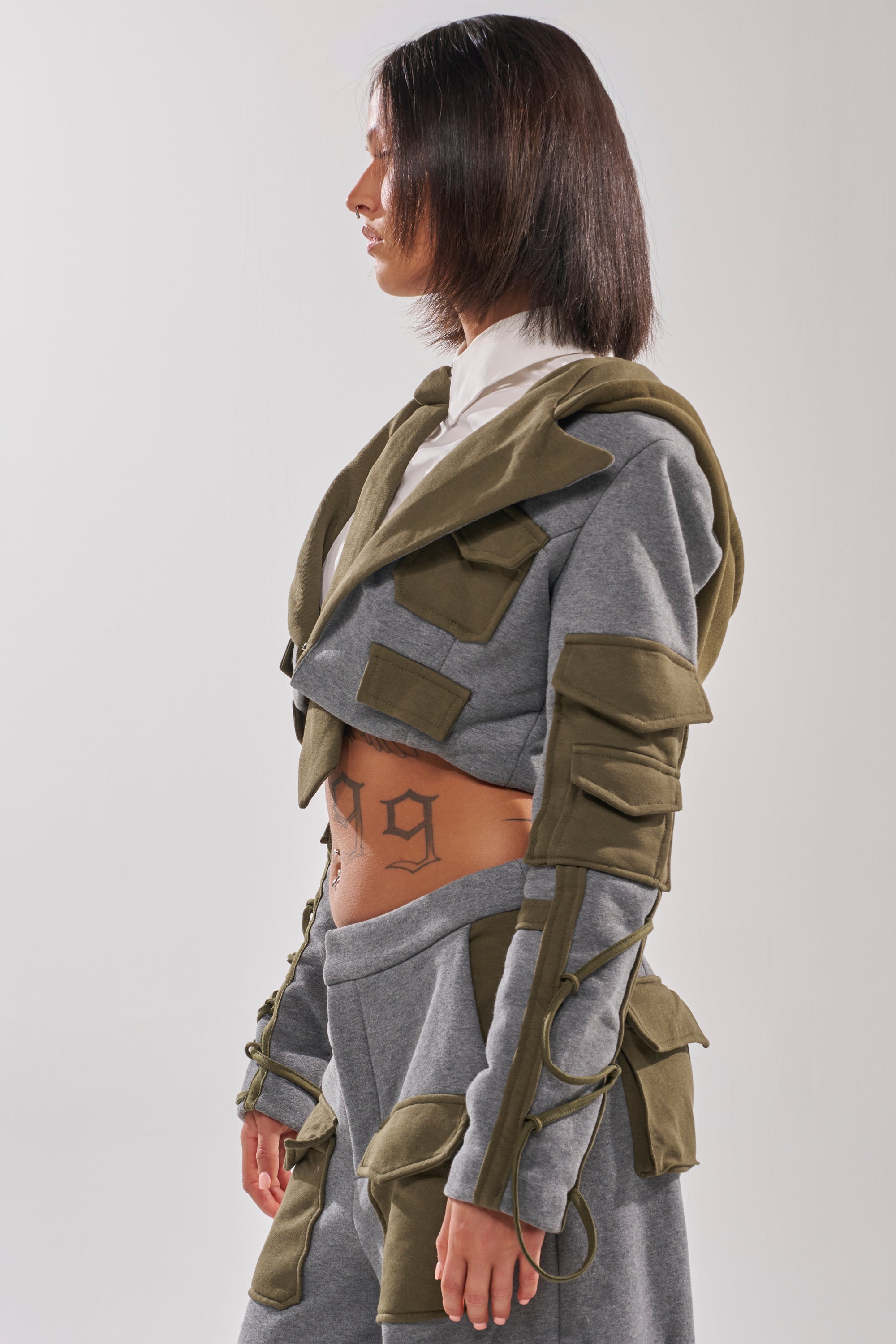 A person with straight, shoulder-length dark hair wears the CRAZY IN LOVE BLOUSE in gray and olive-green, featuring utility pockets and midriff cutouts. They have a "1999" tattoo on their torso. The background is plain and light-colored.