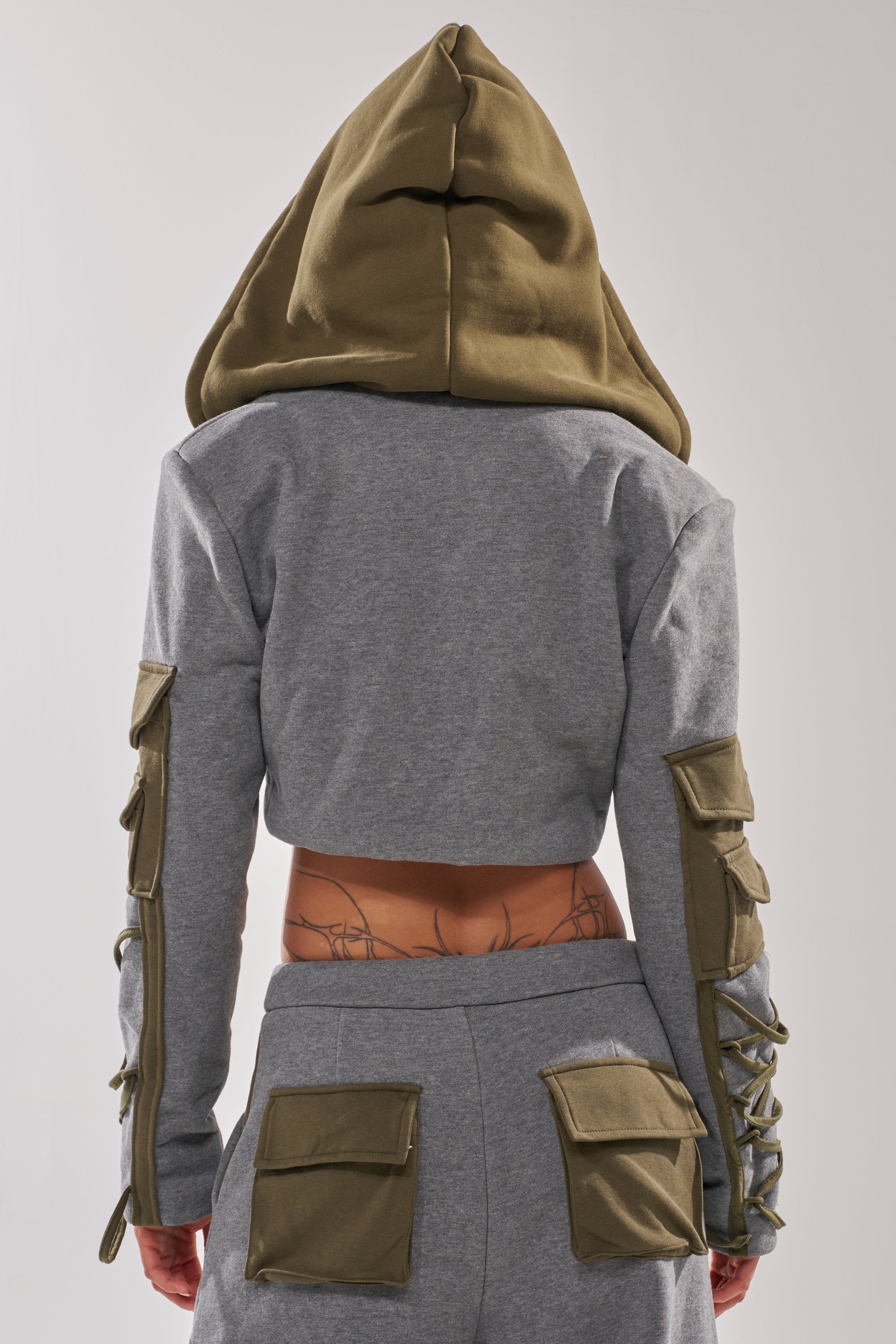 A person wearing the CRAZY IN LOVE BLOUSE—a cropped gray jacket with utility pockets, sleeve laces, and a pointed hood—alongside matching pants with green pockets. The photo is taken from behind, showing part of a lower back tattoo.