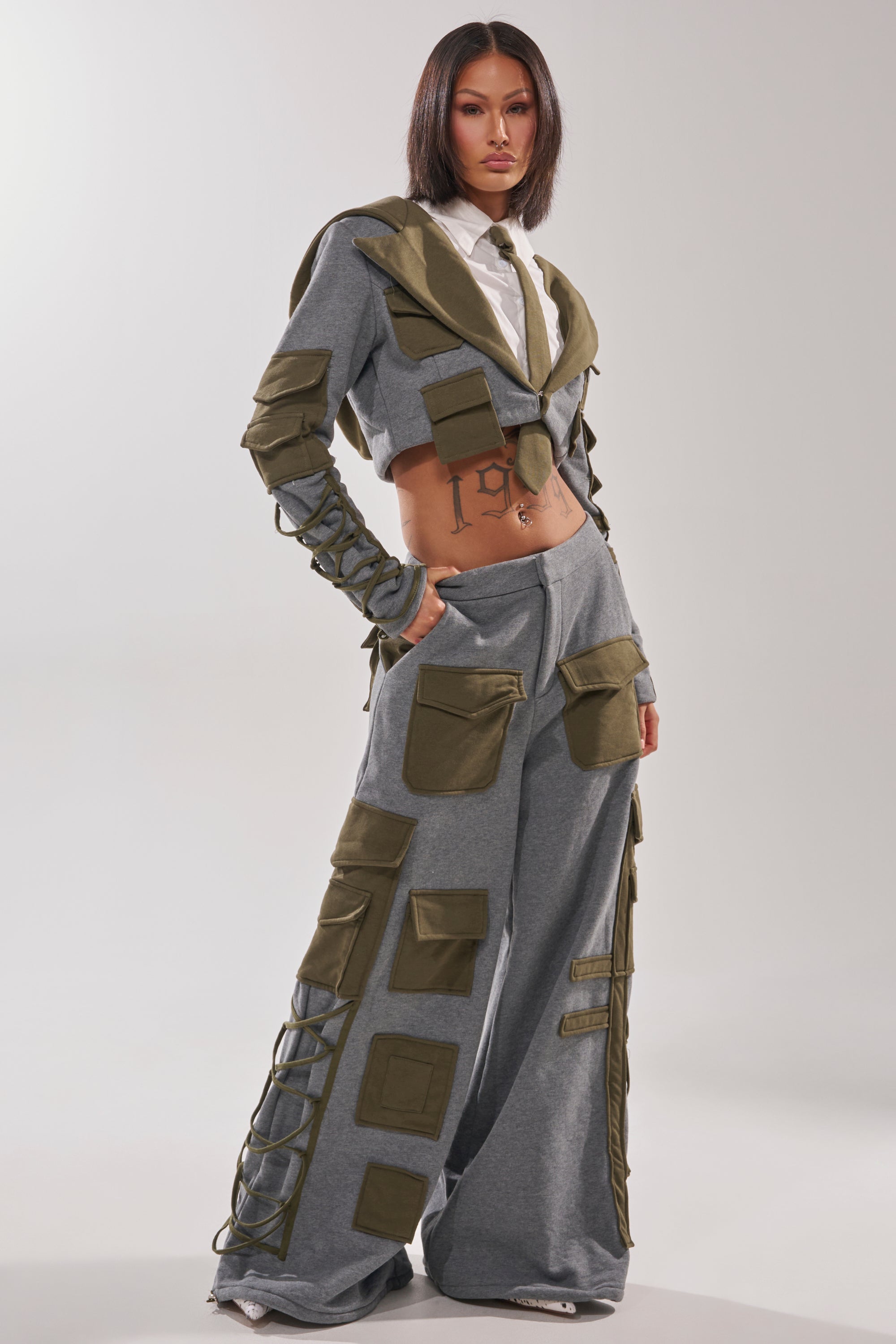 A woman poses in a modern outfit featuring the CRAZY IN LOVE BLOUSE, paired with a cropped grey jacket with olive green accents and matching wide-leg utility pants, revealing her midriff for an edgy streetwear look.