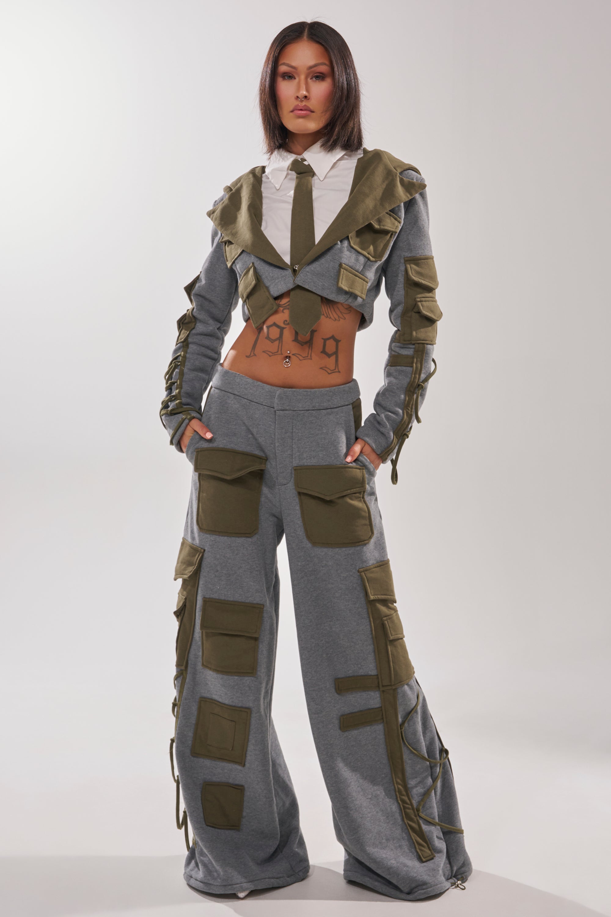 A woman faces forward wearing the CRAZY IN LOVE BLOUSE, a gray streetwear piece with olive green utility pockets and details, styled with wide-leg pants and a cropped jacket over a white shirt and tie. Her exposed midriff reveals a large tattoo.