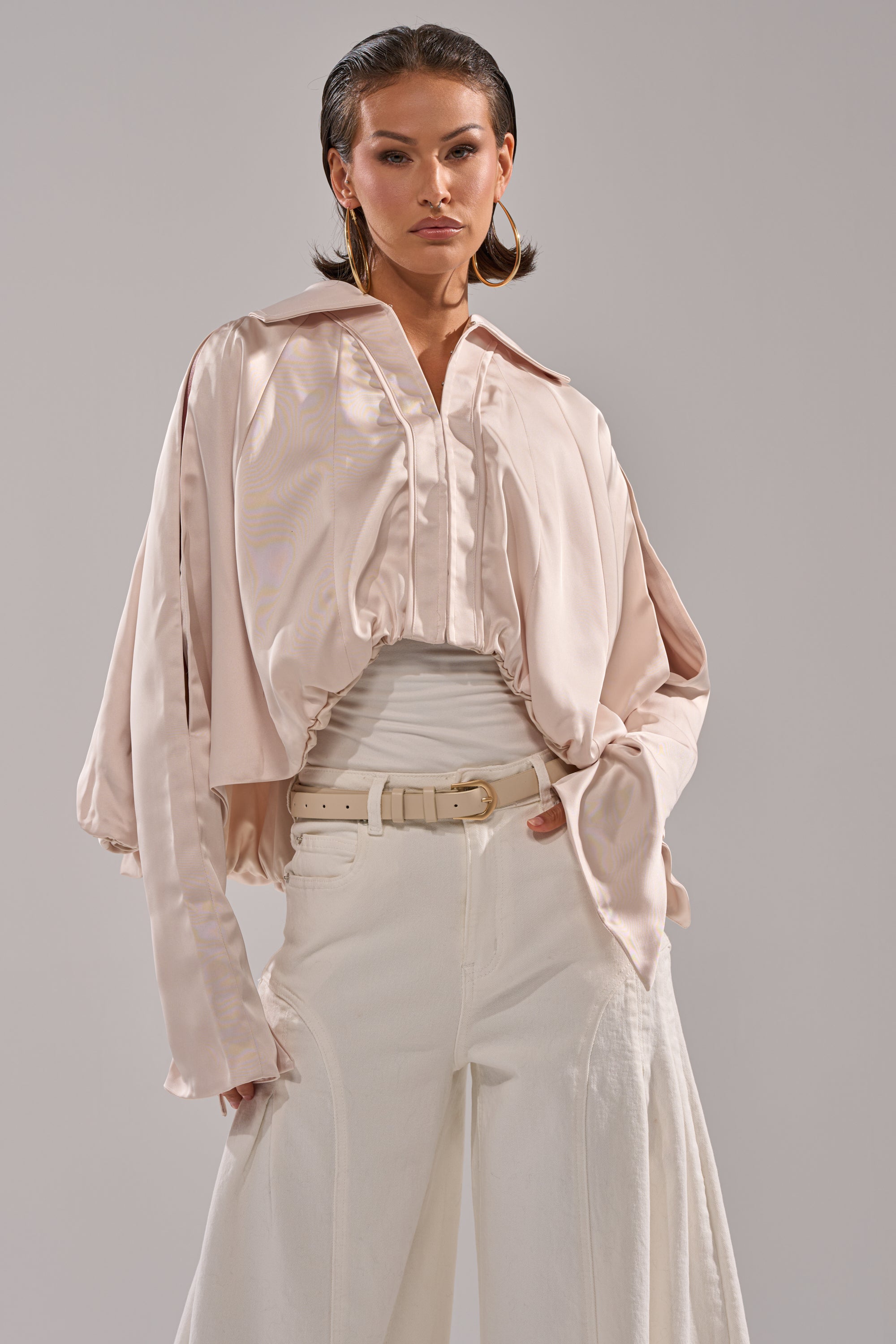 A woman poses confidently in the GET WITH IT SATIN BLOUSE—loose, pale pink with dolman sleeves—layered over a white top and paired with wide-leg white pants, a matching belt, slicked-back hair, and large hoop earrings. 0