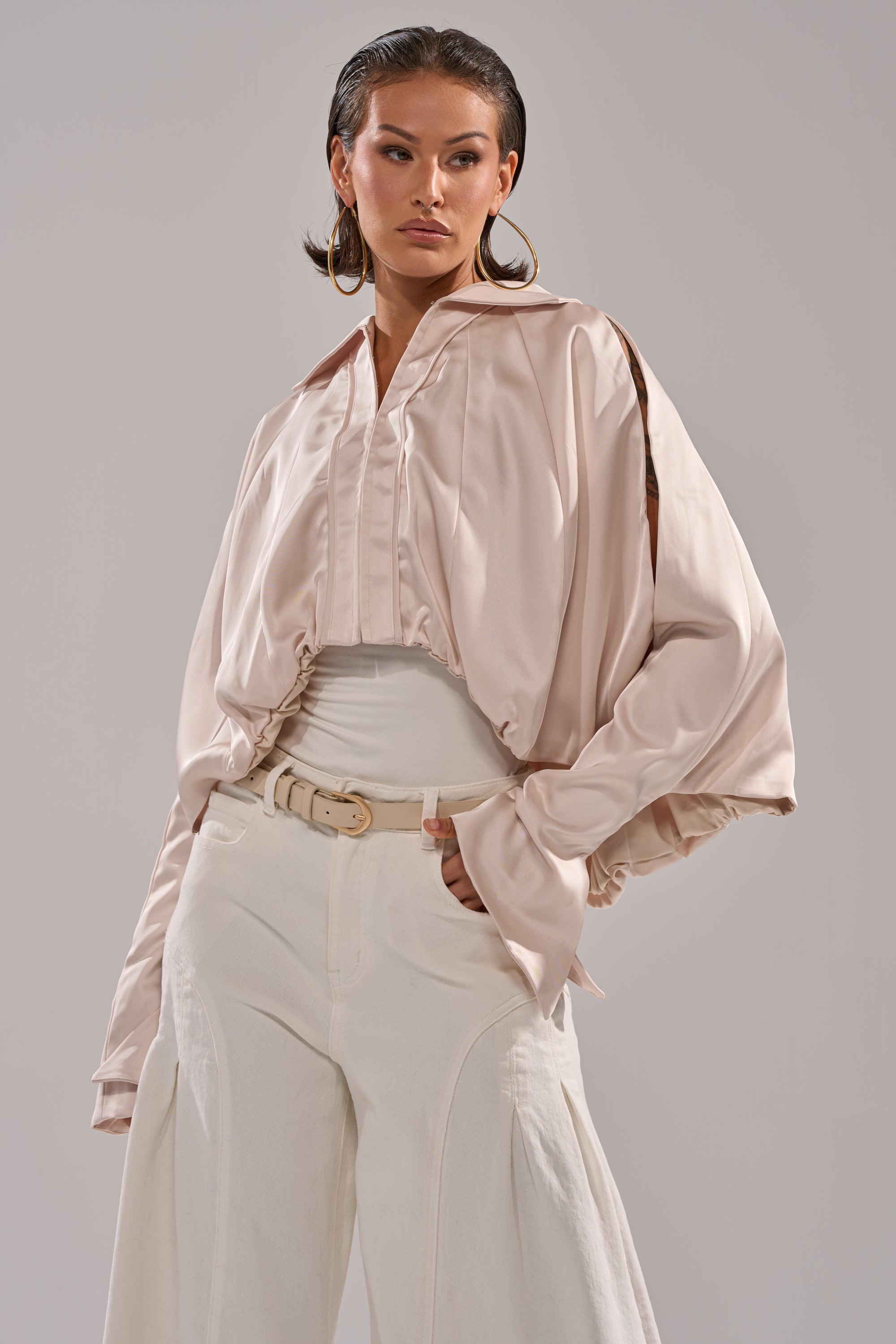 A woman exudes confidence in the GET WITH IT SATIN BLOUSE, a shiny light pink top with dolman sleeves, styled with a white belt, wide-leg white pants, slicked-back hair, and large hoop earrings against a plain background. 1
