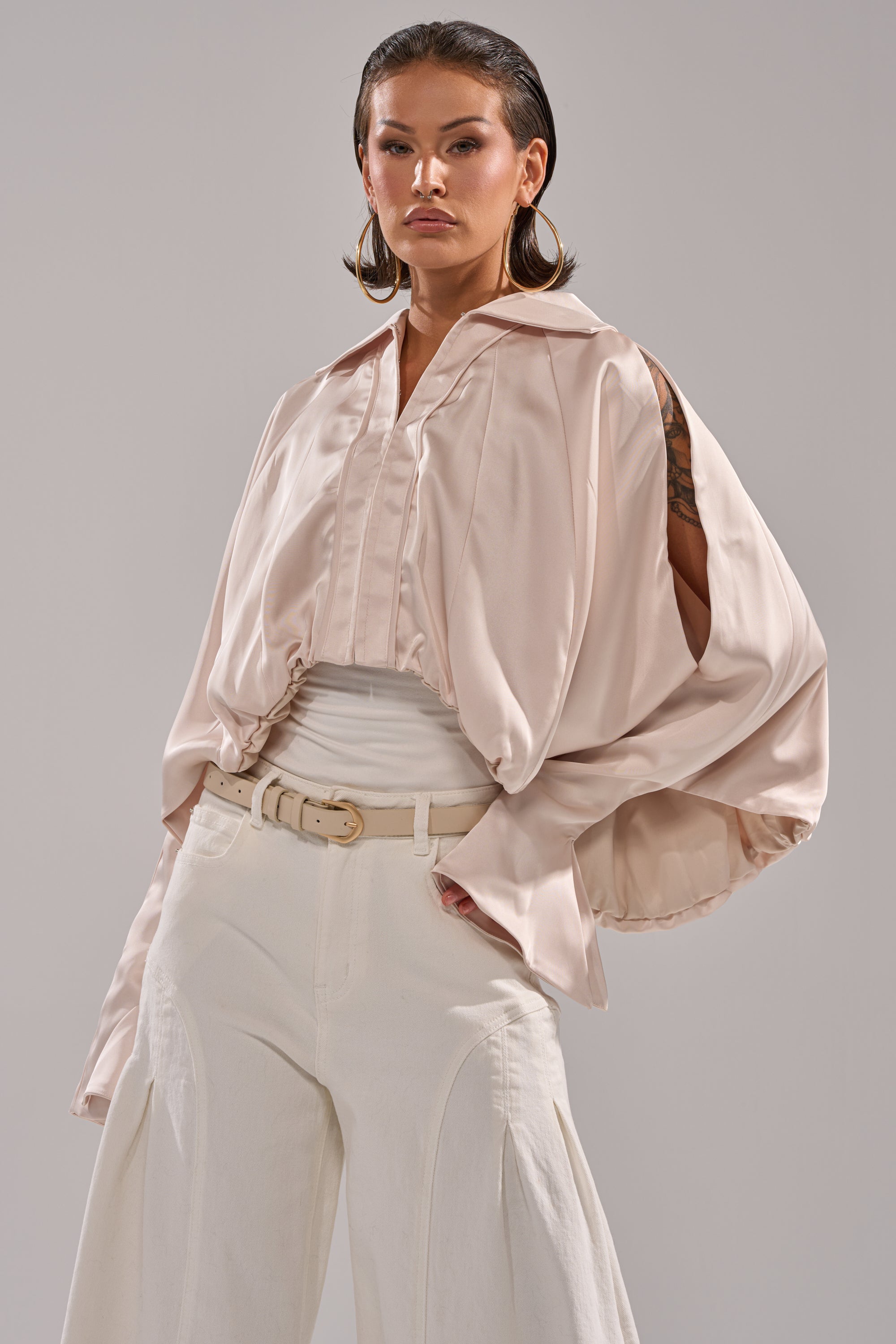 A person stands confidently in the GET WITH IT SATIN BLOUSE—light beige with dolman sleeves and cutout shoulders—paired with high-waisted white pants and hoop earrings, set against a plain gray background. 2