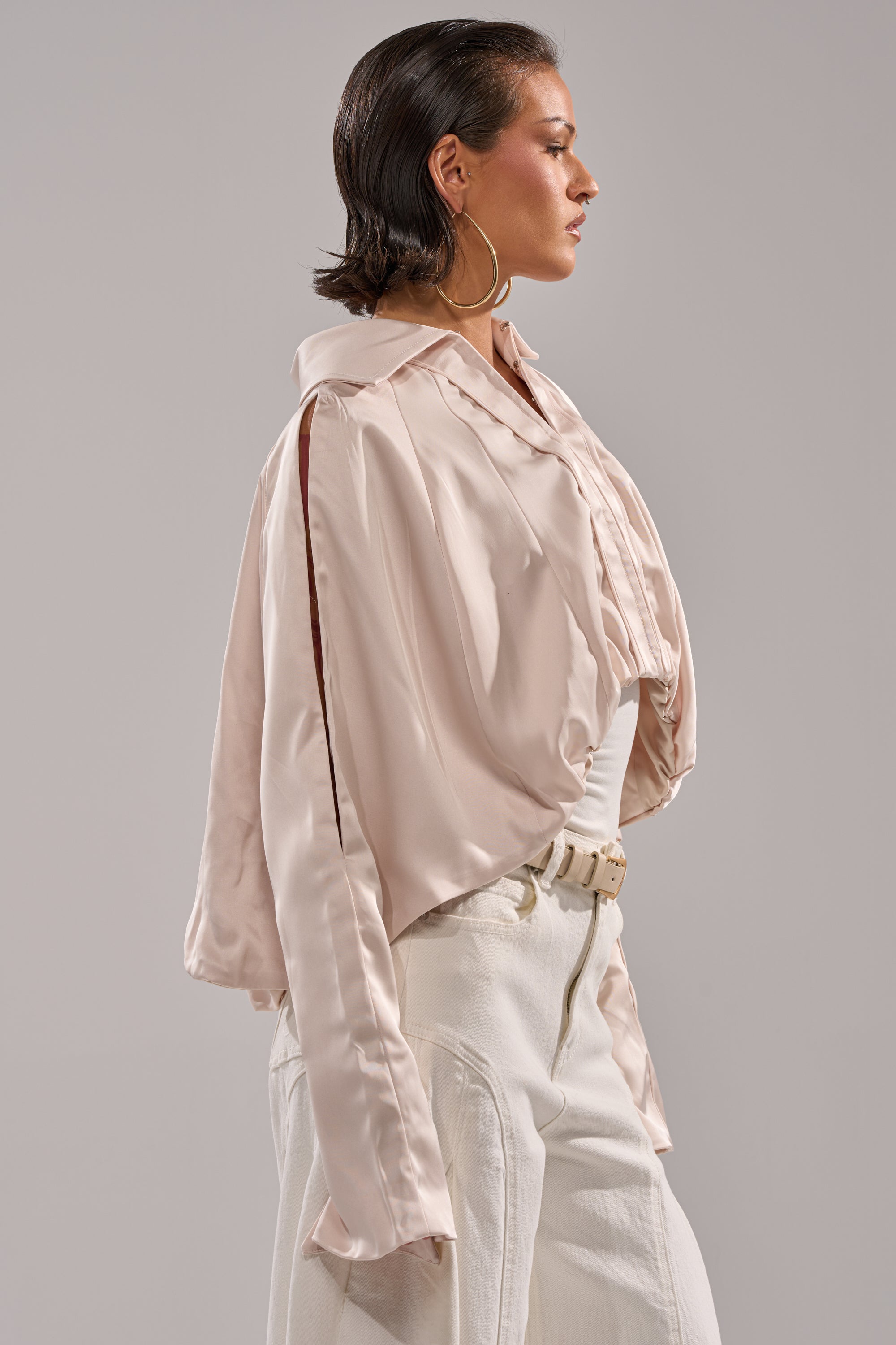 A woman with slicked-back hair wears the GET WITH IT SATIN BLOUSE, a shiny light pink cropped top with wide dolman sleeves, paired with white high-waisted pants, large hoop earrings, and a white belt against a plain background. 4