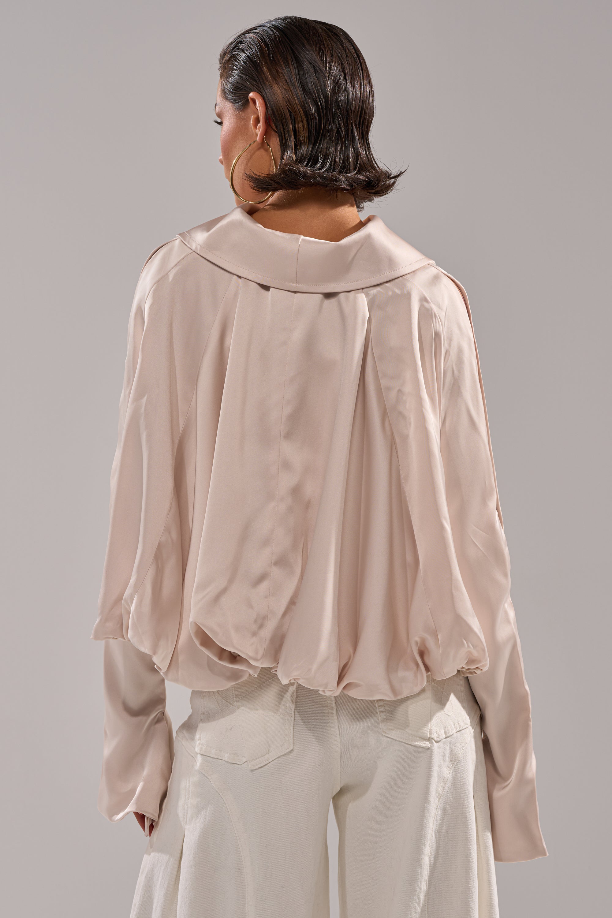 A woman with short, slicked-back hair is seen from behind wearing the GET WITH IT SATIN BLOUSE—a loose, light beige top with wide Dolman sleeves, pleated accents, and a soft sheen—paired with white pants against a plain gray background. 5