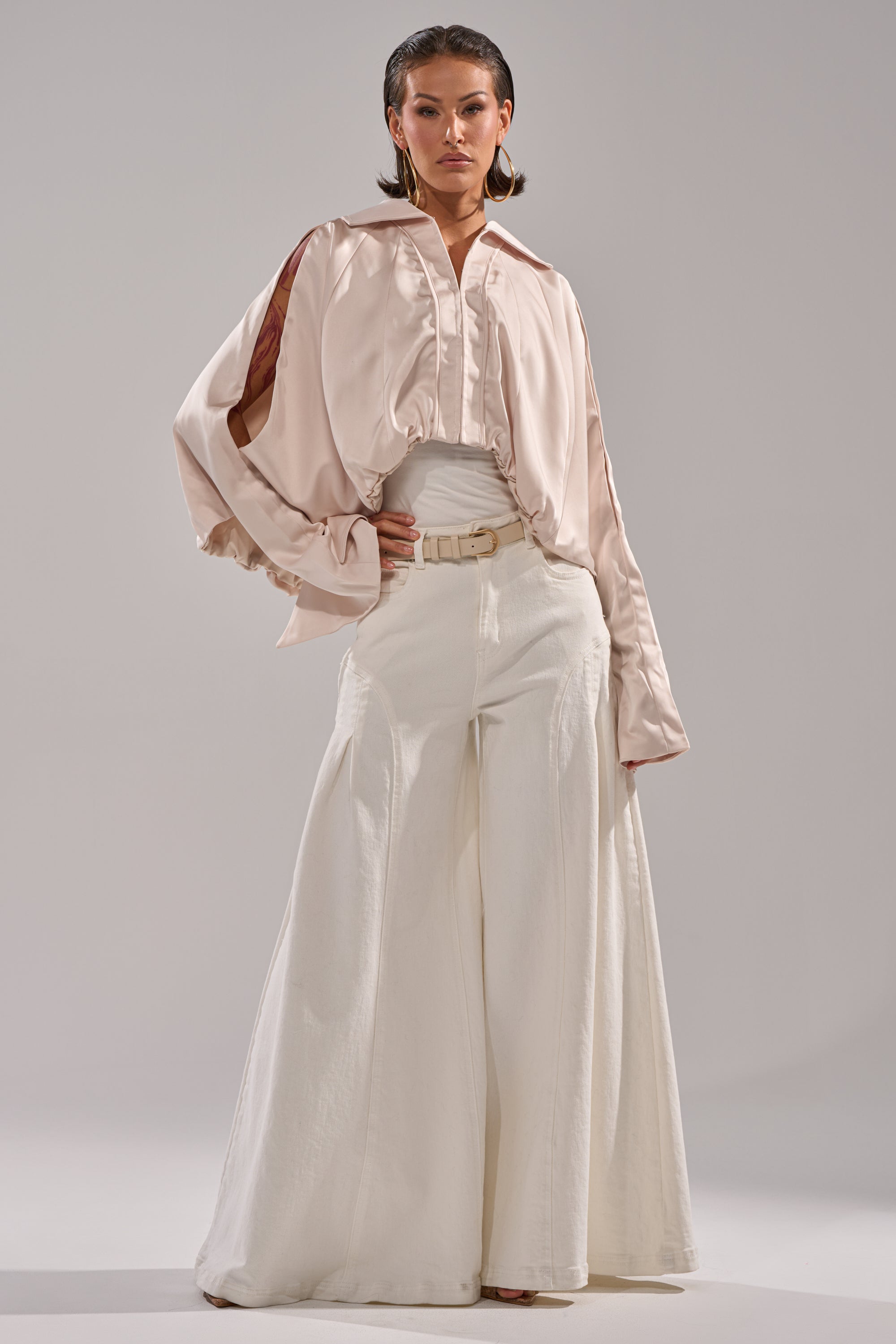 A person stands confidently in the GET WITH IT SATIN BLOUSE—light beige with Dolman sleeves—paired with high-waisted, wide-leg white pants, a belt, and large hoop earrings against a plain grey background. 7