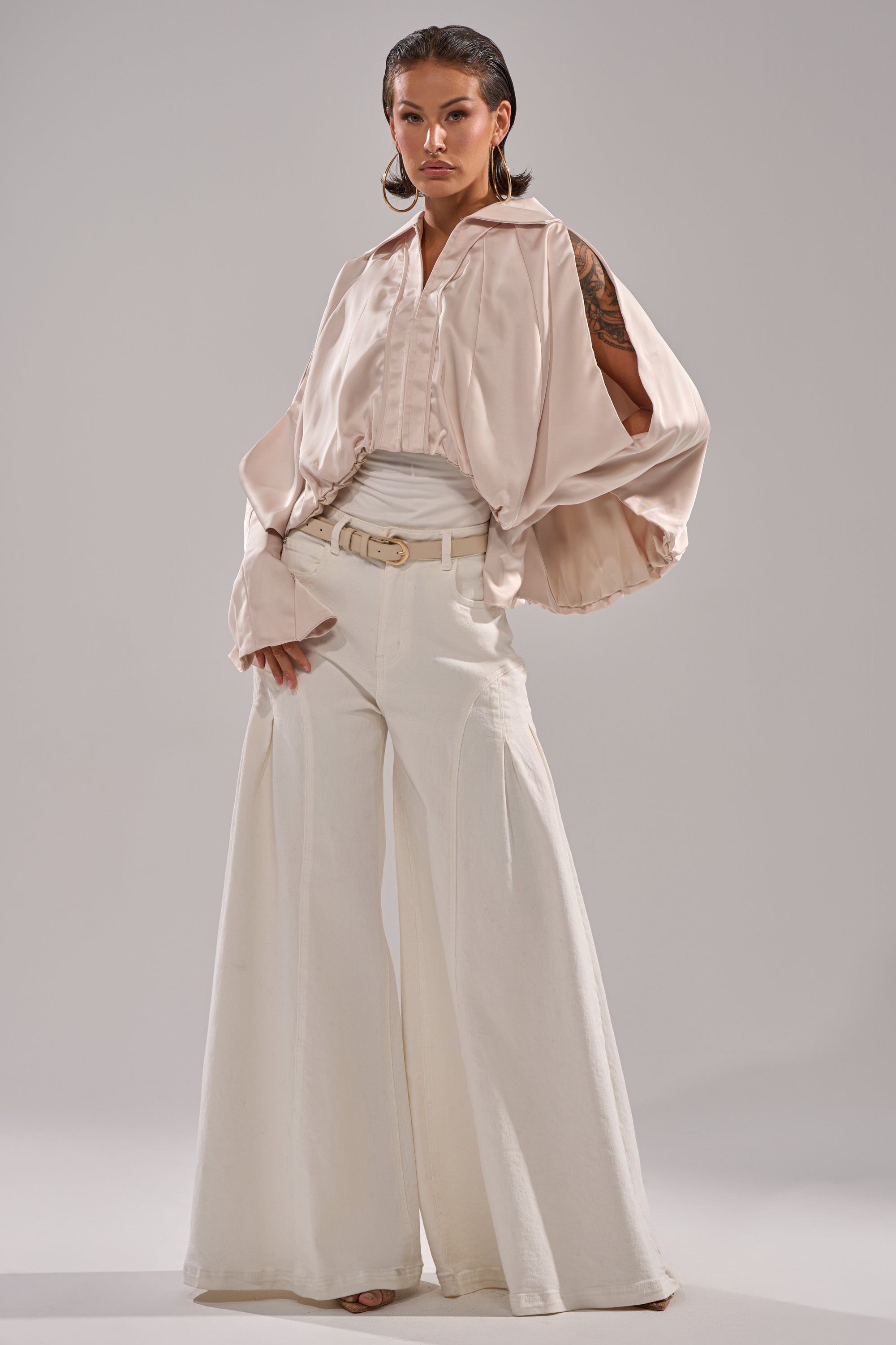 A woman exudes confidence in the GET WITH IT SATIN BLOUSE, a pale pink top with wide Dolman sleeves, paired with high-waisted, wide-leg white pants as she poses against a simple light background. 8