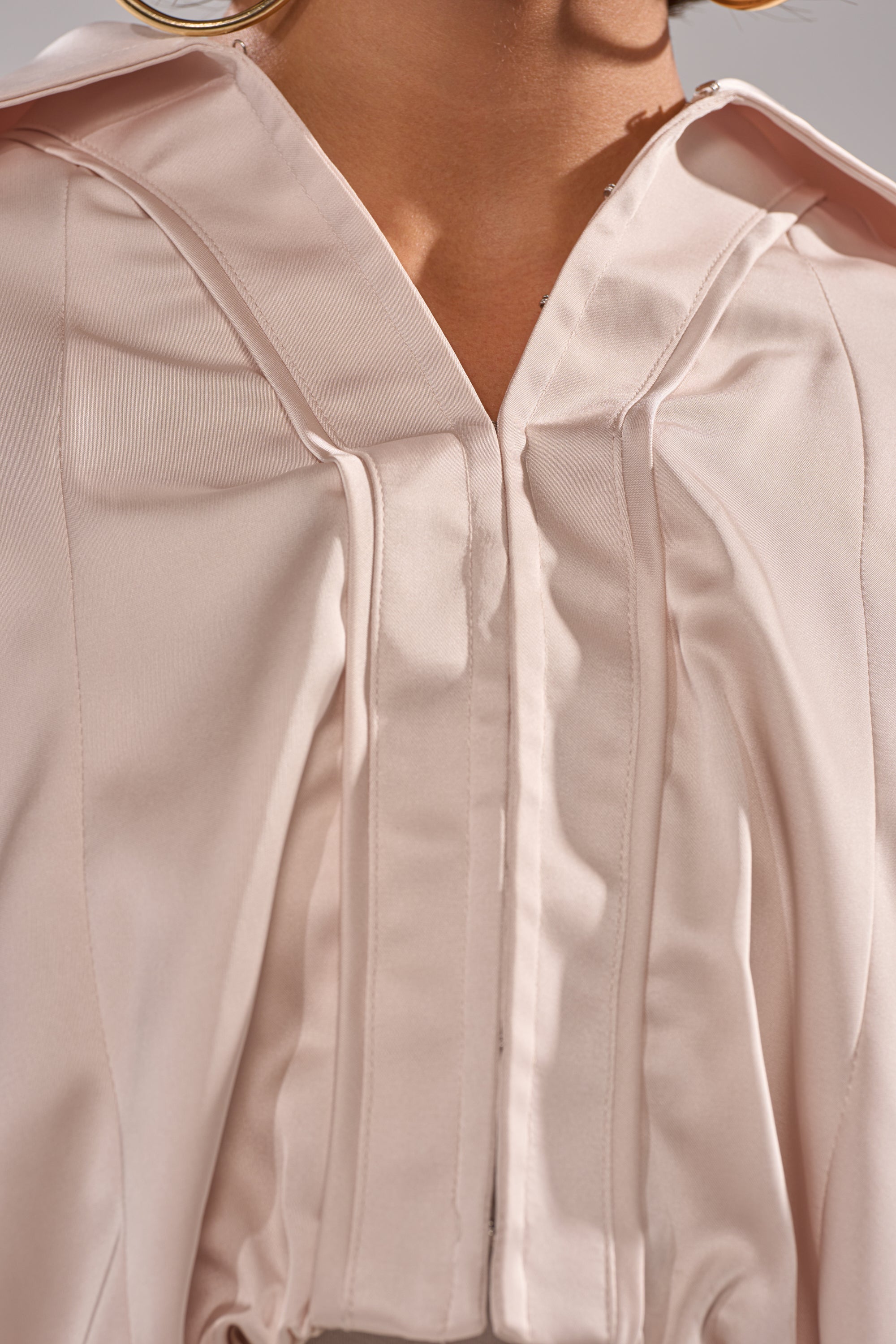 Close-up of a person wearing the GET WITH IT SATIN BLOUSE in pale pink, featuring dolman sleeves, pleated details, a v-shaped neckline, and a structured collar. The silky fabric catches the light, highlighting its elegant texture. 9