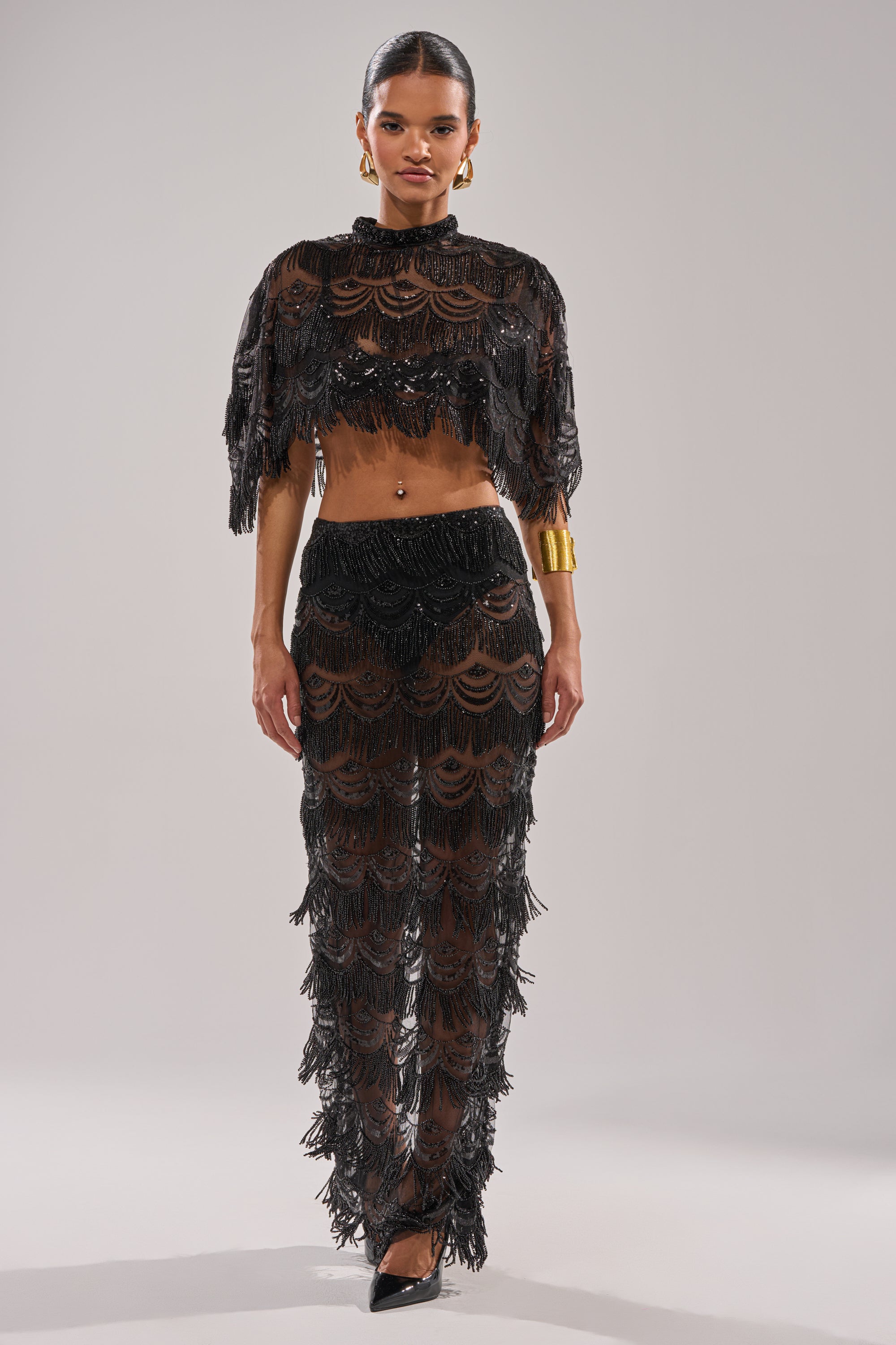 A woman faces forward in the STAR OF THE NIGHT MAXI SKIRT, a high-rise fit maxi with mesh beadwork and fringe. She pairs it with a matching sheer black crop top, gold hoop earrings, a gold cuff bracelet, and black pointed heels.
