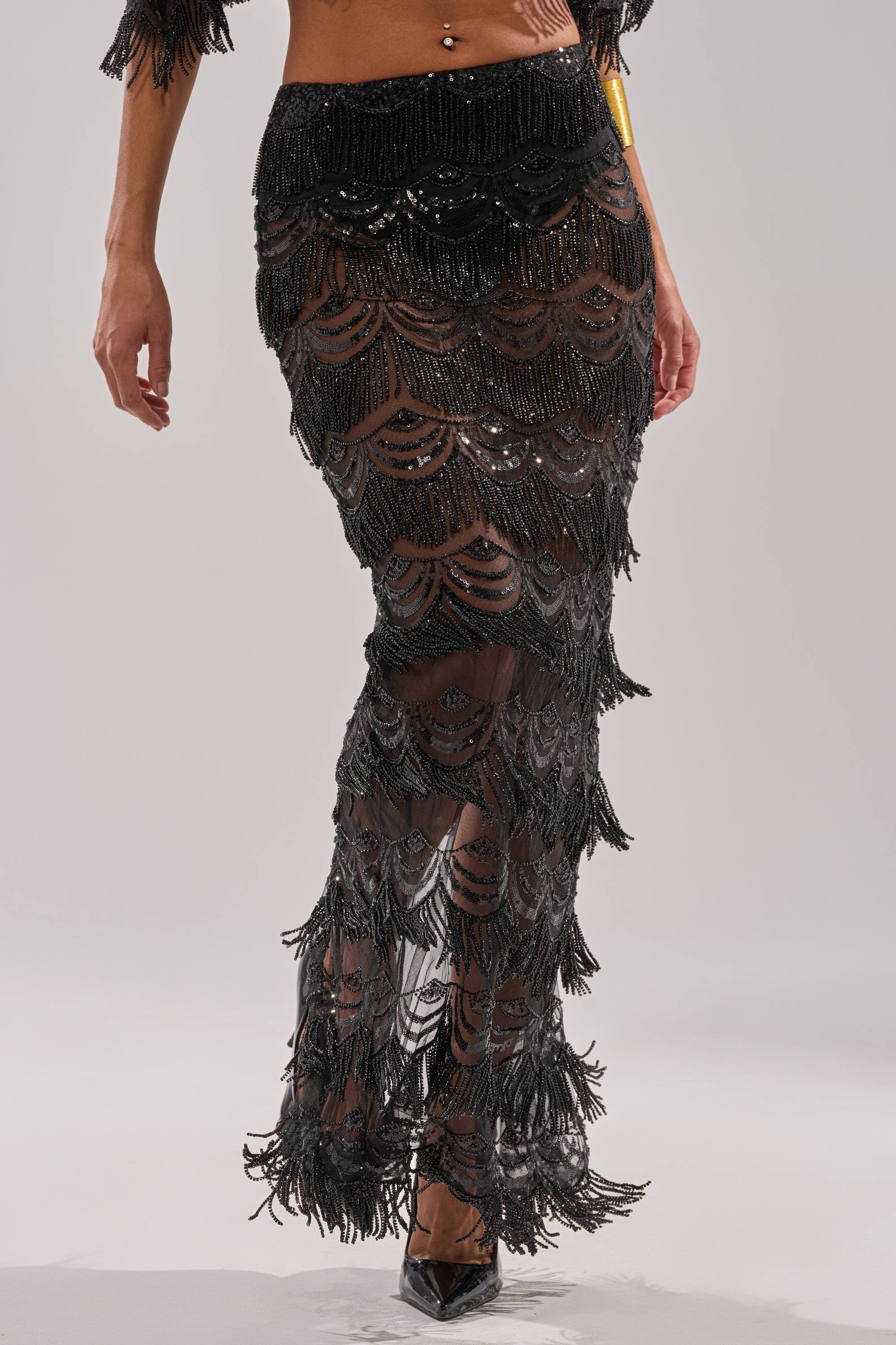 Wearing the STAR OF THE NIGHT MAXI SKIRT—sheer black with fringe accents and scallop patterns—a person reveals their legs, pairing it with black pointed-toe heels. Only the lower half of their body is visible.