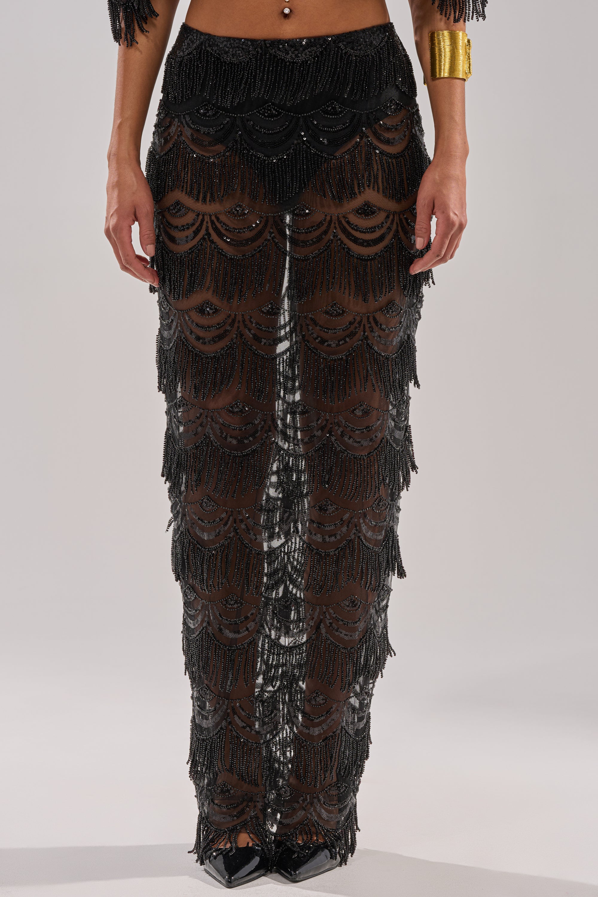 Wearing the STAR OF THE NIGHT MAXI SKIRT featuring black beaded mesh and scalloped edges, paired with black pointed shoes, a gold cuff bracelet, and a belly button piercing, the person stands against a plain background.