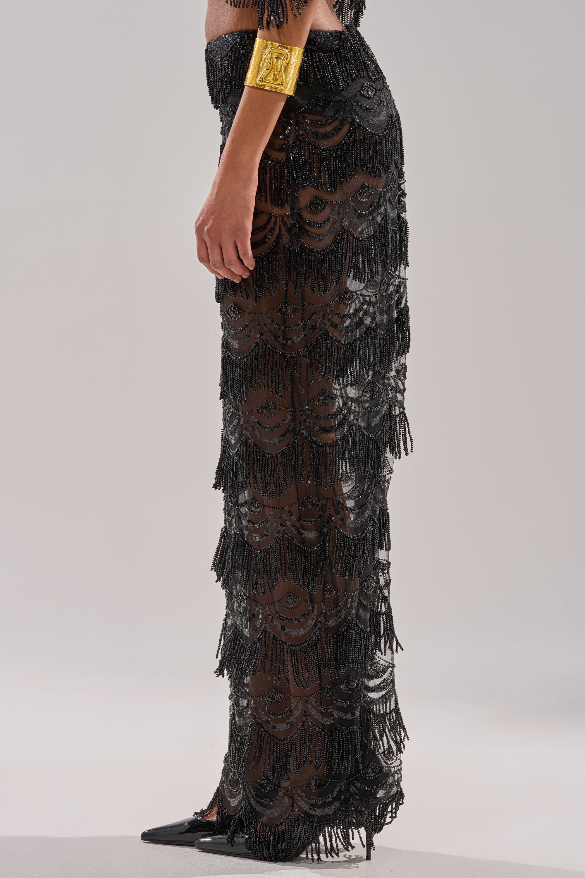 Wearing the STAR OF THE NIGHT MAXI SKIRT, a sheer black beaded mesh design with fringe details, paired with a metallic gold cuff bracelet and black pointed-toe shoes, standing against a plain light background.