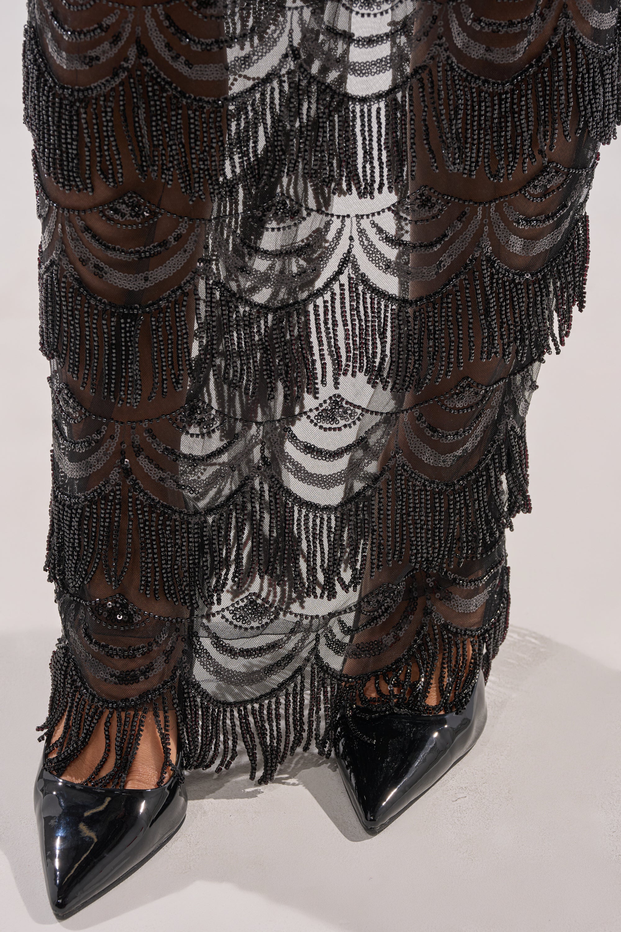 Close-up of a person wearing the STAR OF THE NIGHT MAXI SKIRT, featuring black mesh beadwork with scalloped patterns and beaded fringe, styled with shiny black pointed-toe shoes on a light-colored floor.