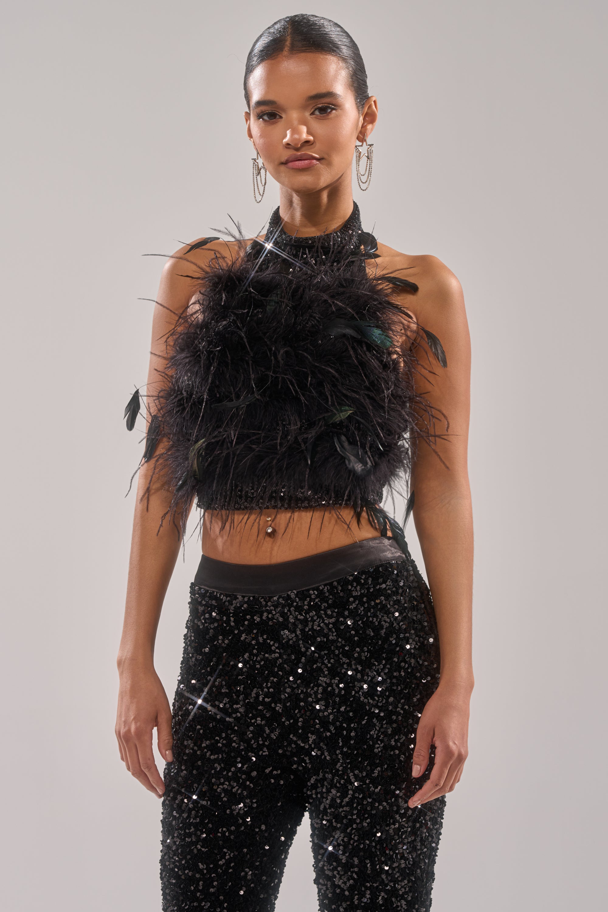 A woman with long dark hair in a ponytail and hoop earrings models the FEATHER BABY BLOUSE, a black cropped feather blouse, paired with black sequined pants against a plain grey background. 0