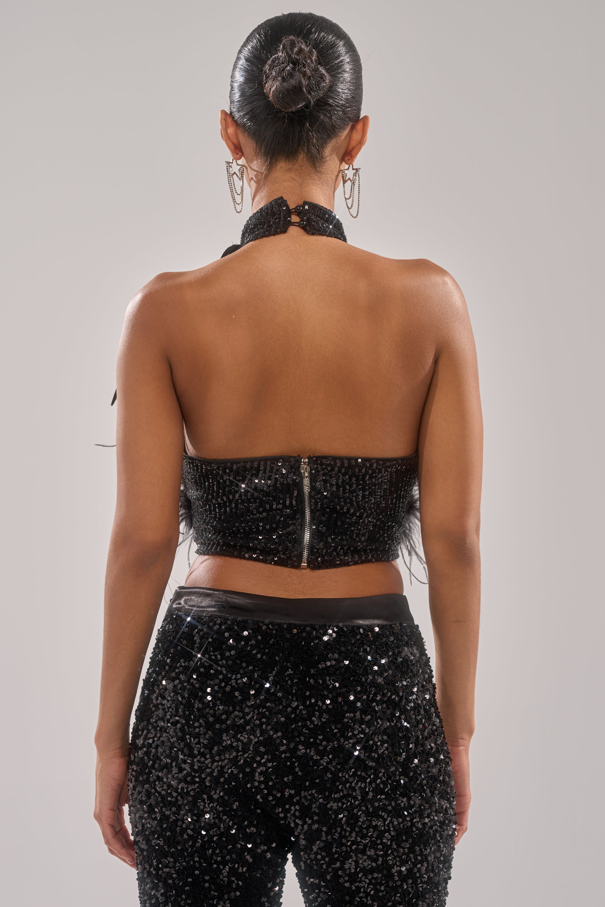 A woman with dark hair in a sleek bun is shown from behind, wearing the FEATHER BABY BLOUSE—a sparkly black sequin top—paired with matching sequin pants against a plain light background. 3
