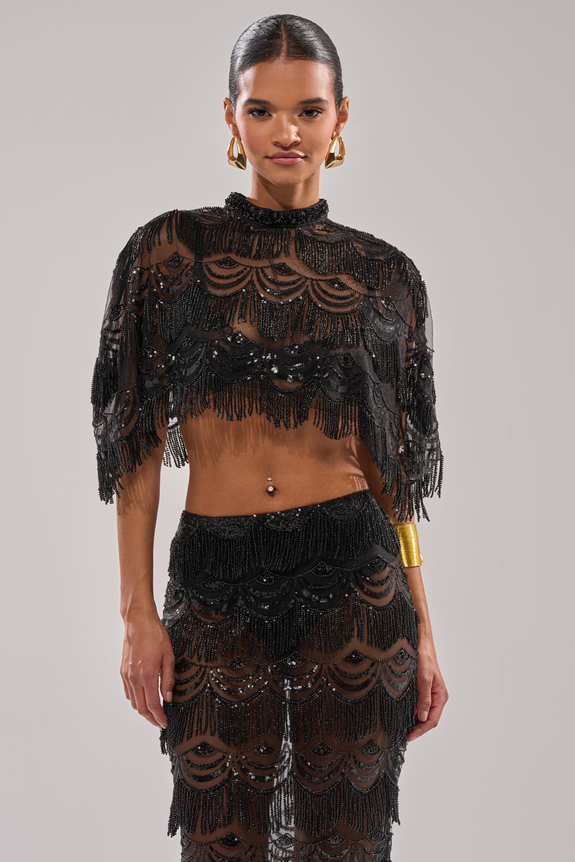 A woman faces forward in the STAR OF THE NIGHT BLOUSE—a sheer black sequin top with scalloped patterns and fringe. She pairs it with gold hoop earrings, a gold cuff bracelet, slicked-back hair, and stands before a light gray background.