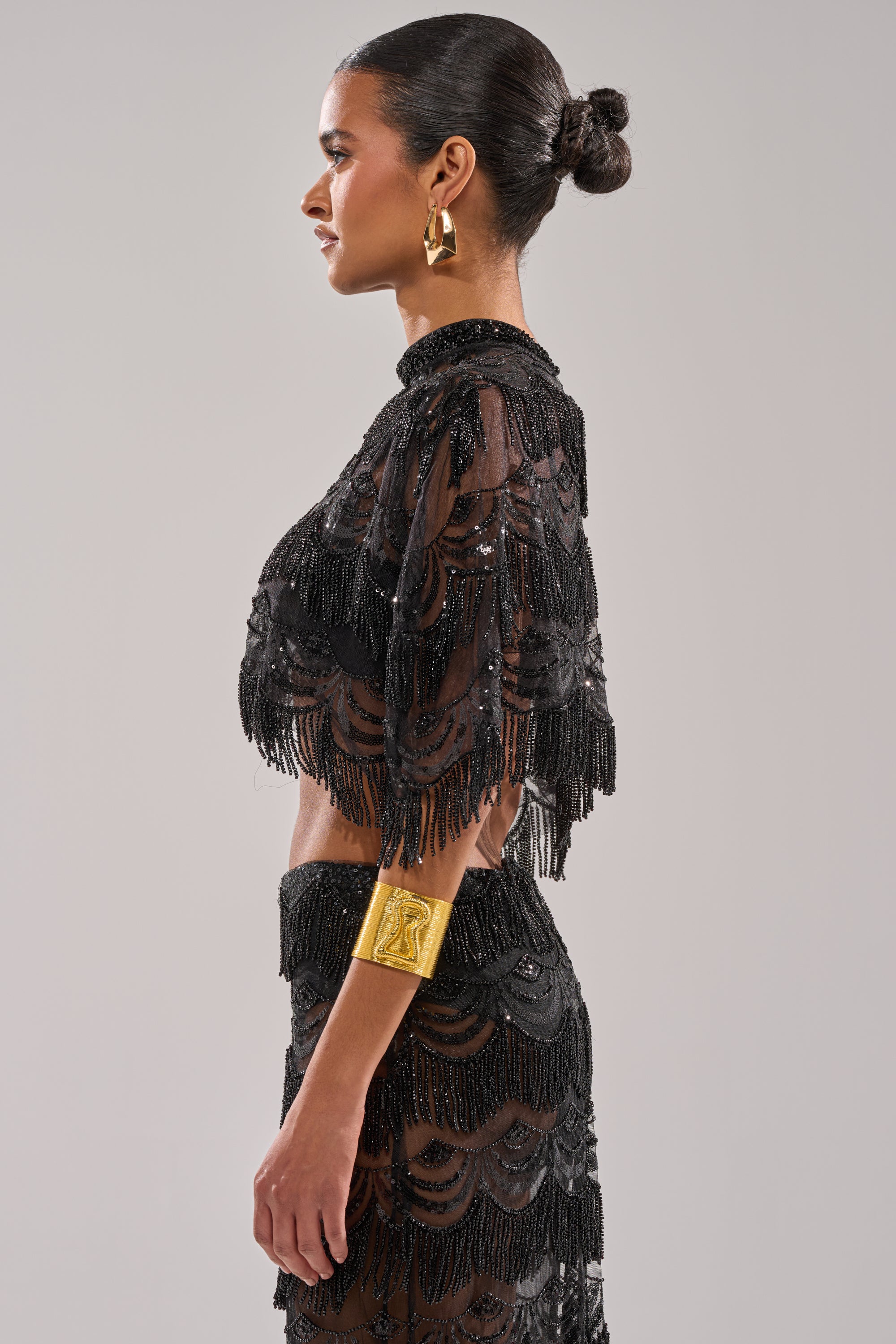 A woman in profile wears the STAR OF THE NIGHT BLOUSE—a black, fringed, semi-sheer beaded top with a matching skirt. Her hair is in a neat bun, accessorized with gold hoops and a wide cuff. The background is plain light gray.