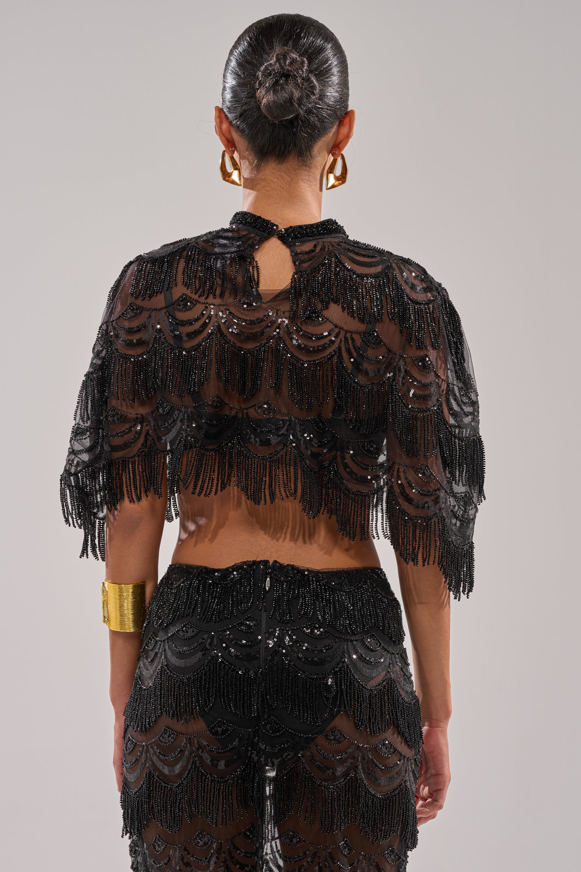 A woman seen from behind wears the STAR OF THE NIGHT BLOUSE, a sheer black beaded top with scalloped patterns, paired with matching pants. Her dark hair is in a sleek bun, accessorized with gold hoop earrings and a wide gold bracelet.