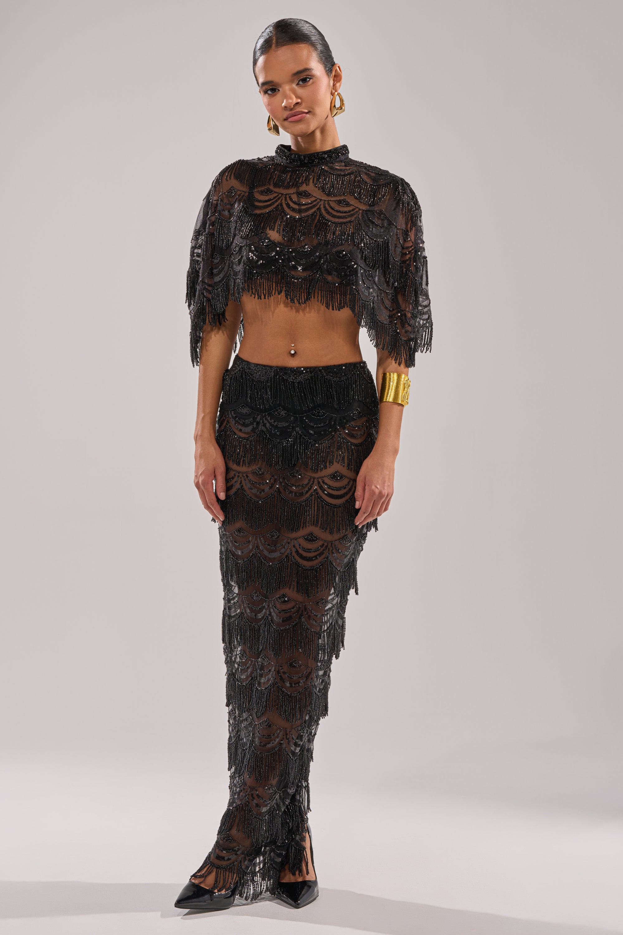 A woman models the STAR OF THE NIGHT BLOUSE, a sheer black lace crop top with beaded fringe, paired with a matching long skirt, black heels, gold hoop earrings, and a wide gold cuff bracelet against a plain light background.