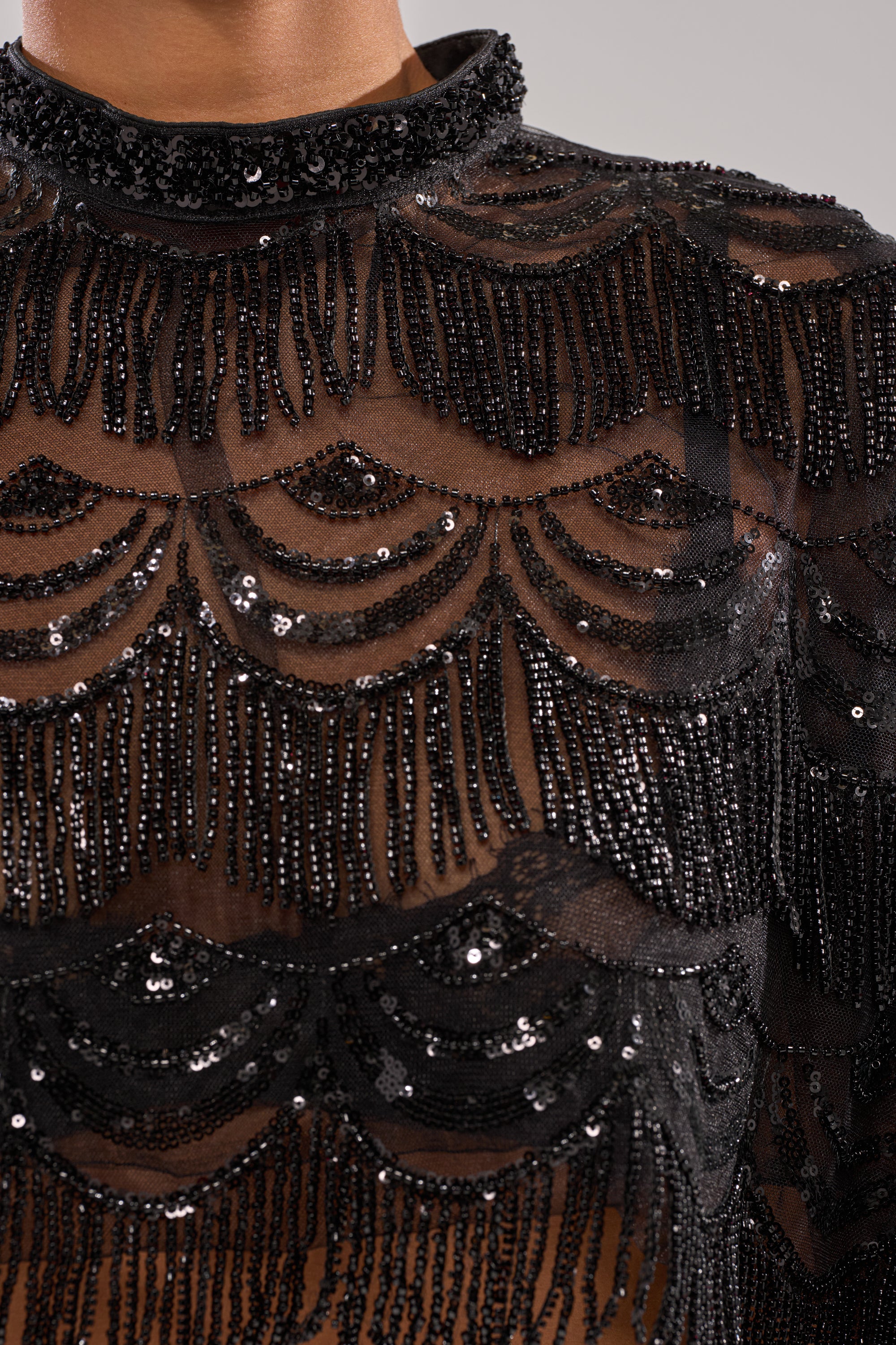 Close-up of the STAR OF THE NIGHT BLOUSE—a sheer black blouse featuring intricate black sequin patterns and beaded fringe in scalloped, wavy designs, styled with a high neckline.
