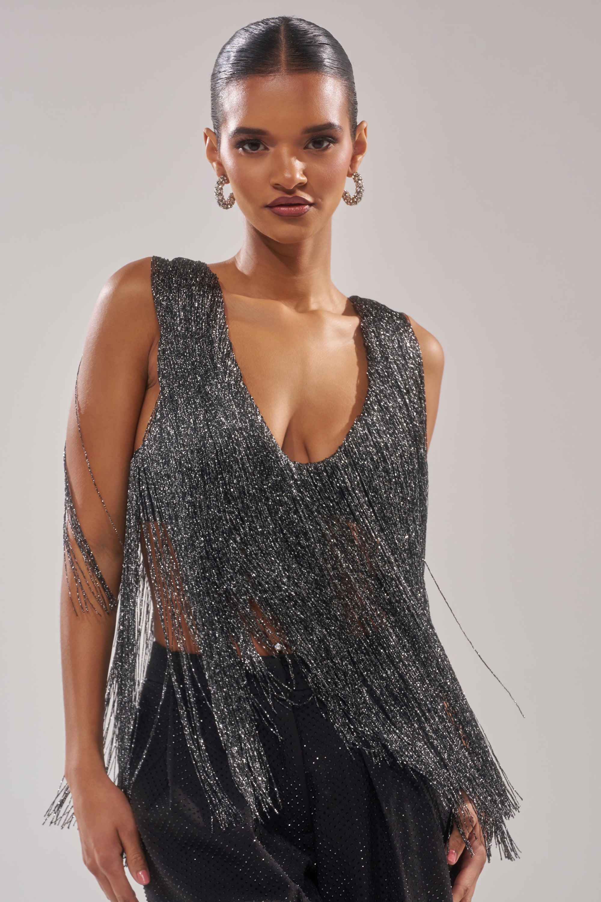 A woman with slicked-back hair confidently poses against a plain light background, wearing the DEVVON FRINGE TOP—a sleeveless, shimmering metallic party top with silver fringe—paired with black pants and large hoop earrings. 1