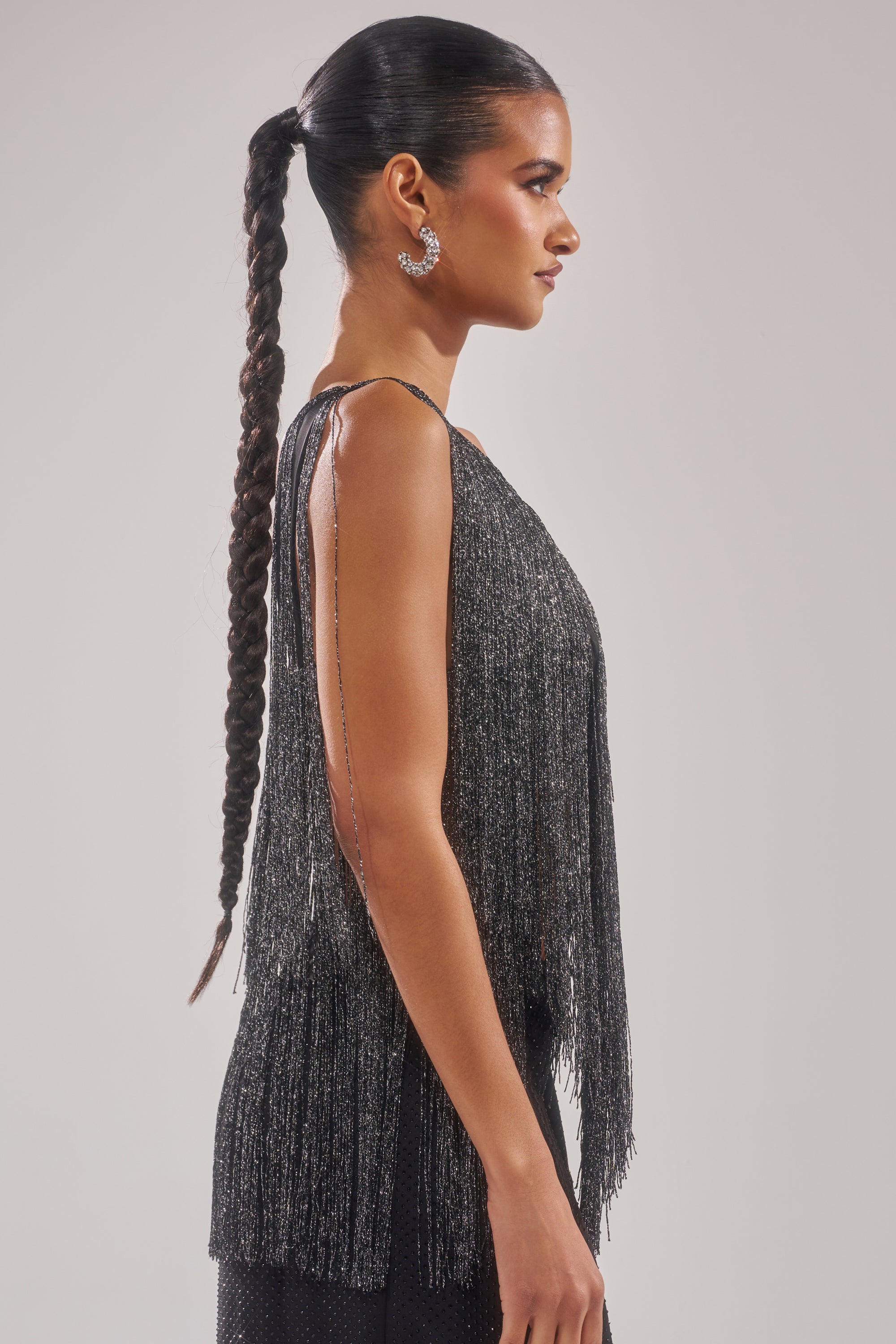 A woman with a long braided ponytail and hoop earrings poses in profile against a plain light background, wearing the shimmery black DEVVON FRINGE TOP woven with metallic thread. 4
