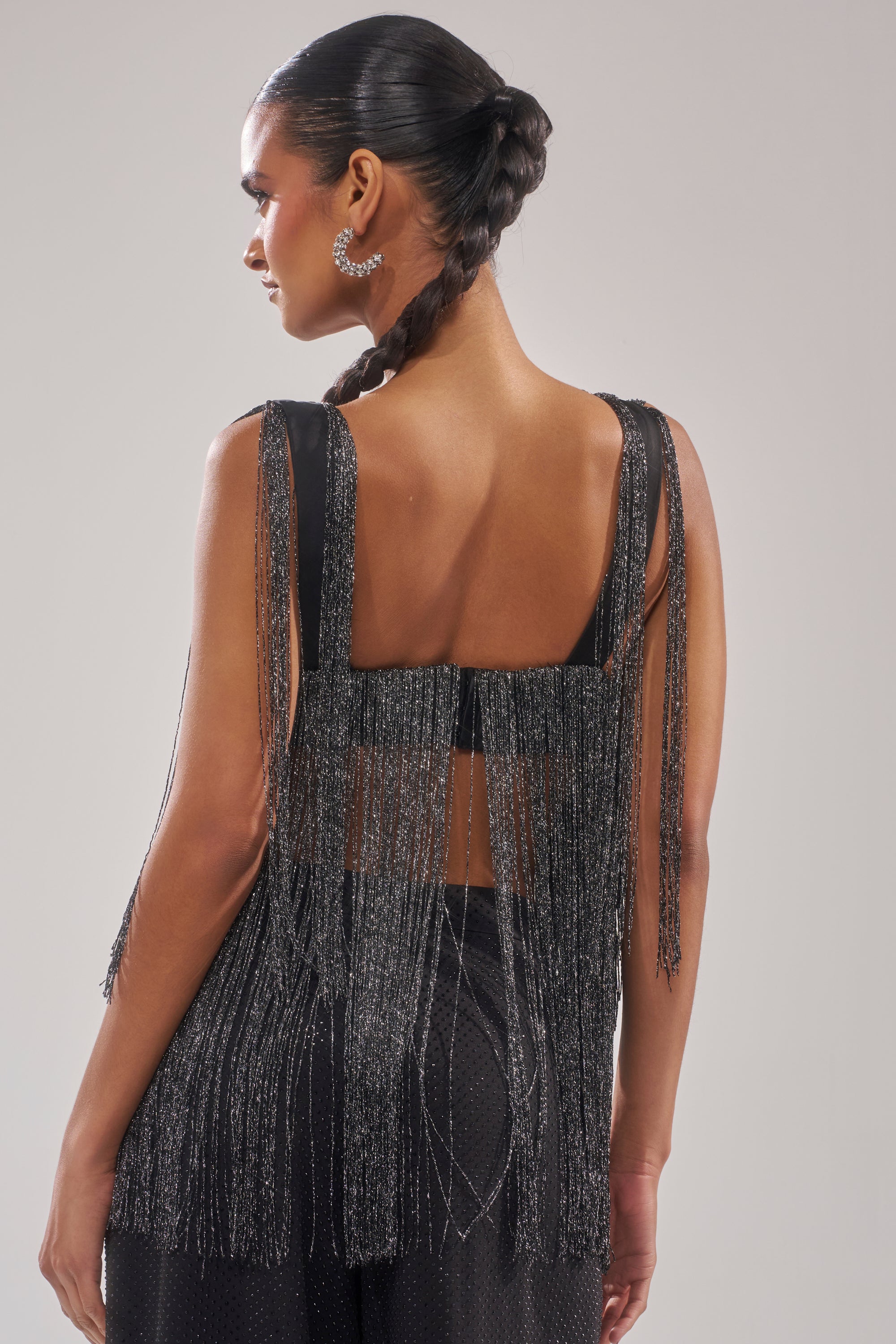 A woman with a sleek braided hairstyle wears the DEVVON FRINGE TOP— a sleeveless black top woven with metallic thread and matching pants. Turned away from the camera, she highlights the shimmering fringe cascading down her back and shoulders. 5