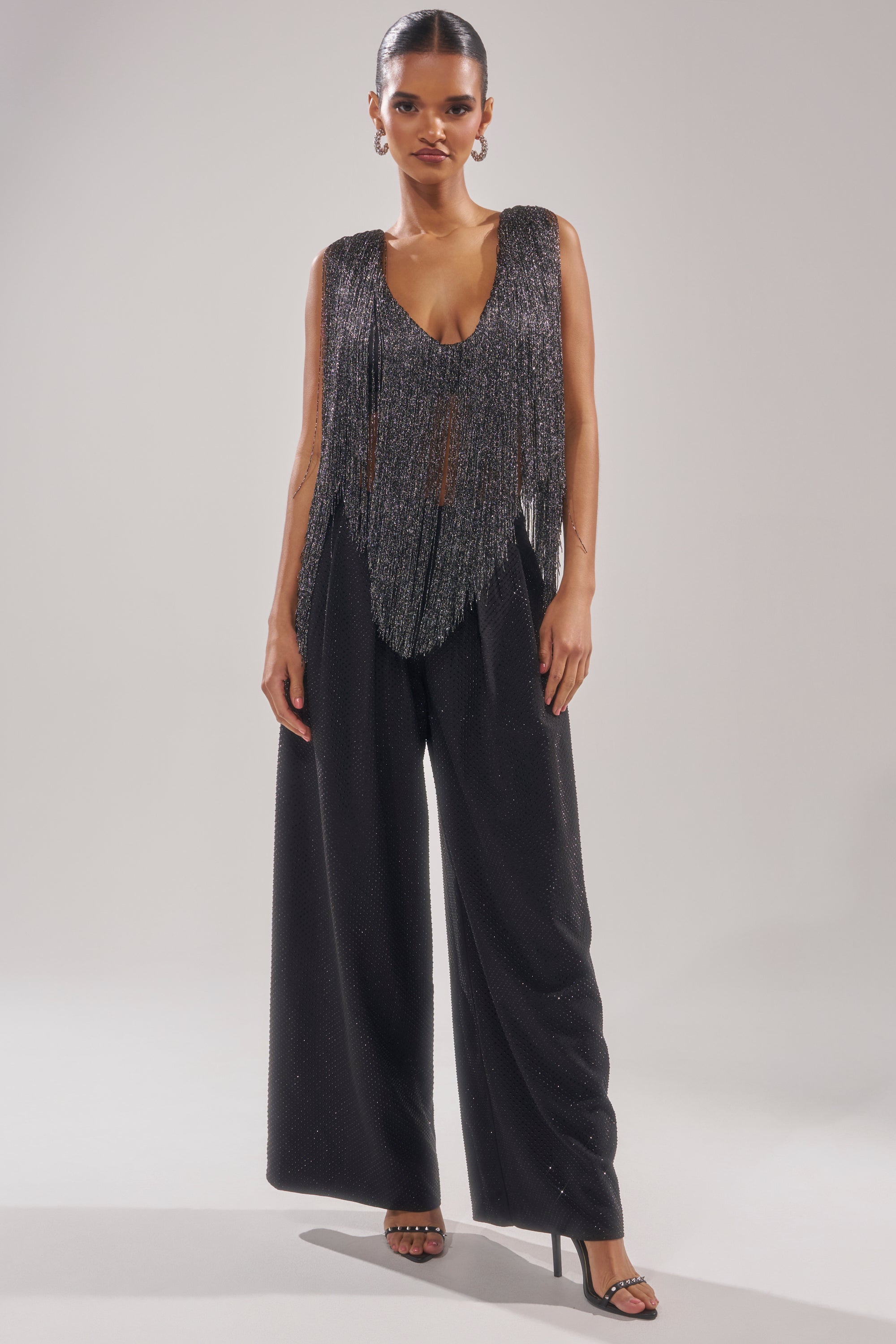 A woman poses against a plain light background wearing the DEVVON FRINGE TOP, a sleeveless sparkly party top with a deep V-neck and silver fringe details, styled with wide-leg black pants and black high-heeled sandals. 6