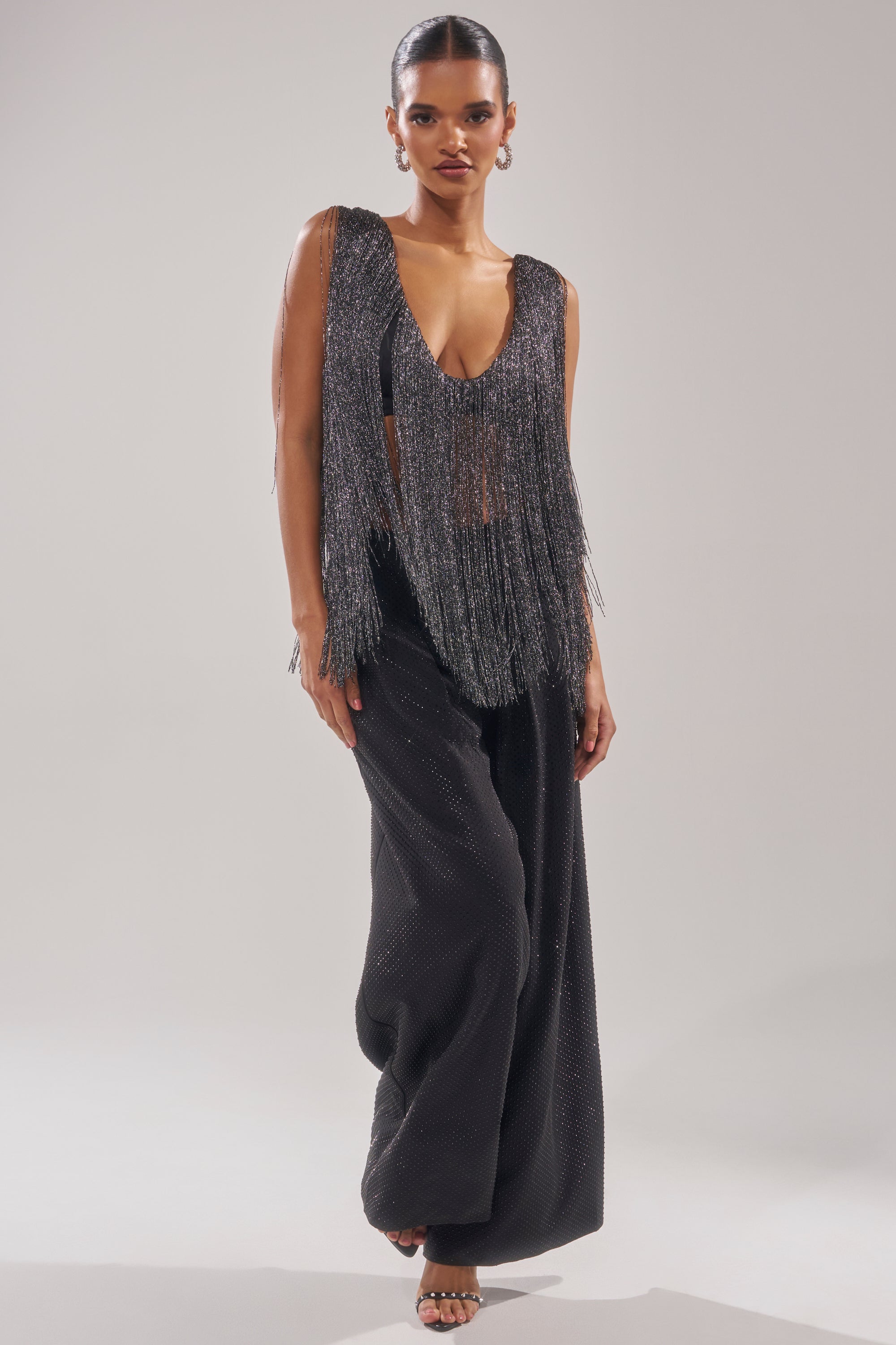 A woman models the DEVVON FRINGE TOP—a sleeveless, deep V-neck with metallic fringe—paired with wide-leg black pants. She stands against a light background, accessorized with hoop earrings and heels for a sparkling party look. 7