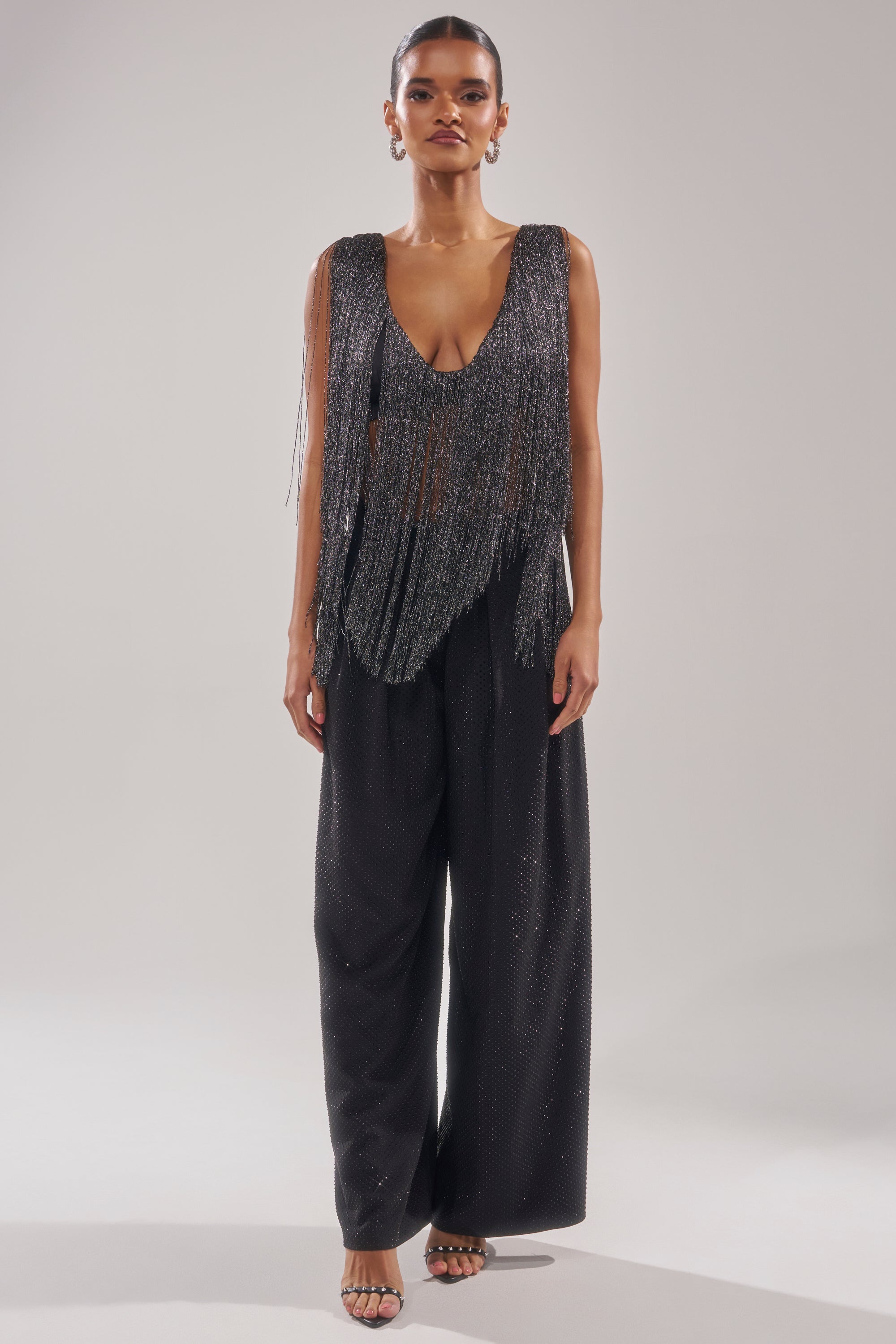 A woman faces forward in the DEVVON FRINGE TOP and wide-leg black pants, her hair pulled back. She accessorizes with hoop earrings and open-toed heels against a plain light background. 8