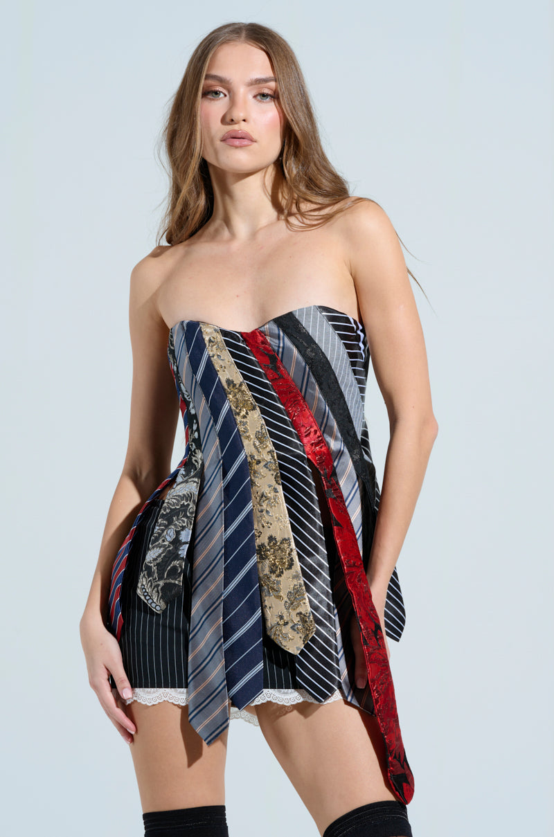A woman with long hair wears the STRICTLY BUSINESS TIE DETAIL CORSET TOP, a unique business-casual dress crafted from diverse neckties in stripes and floral patterns. She stands against a plain, light-colored background.