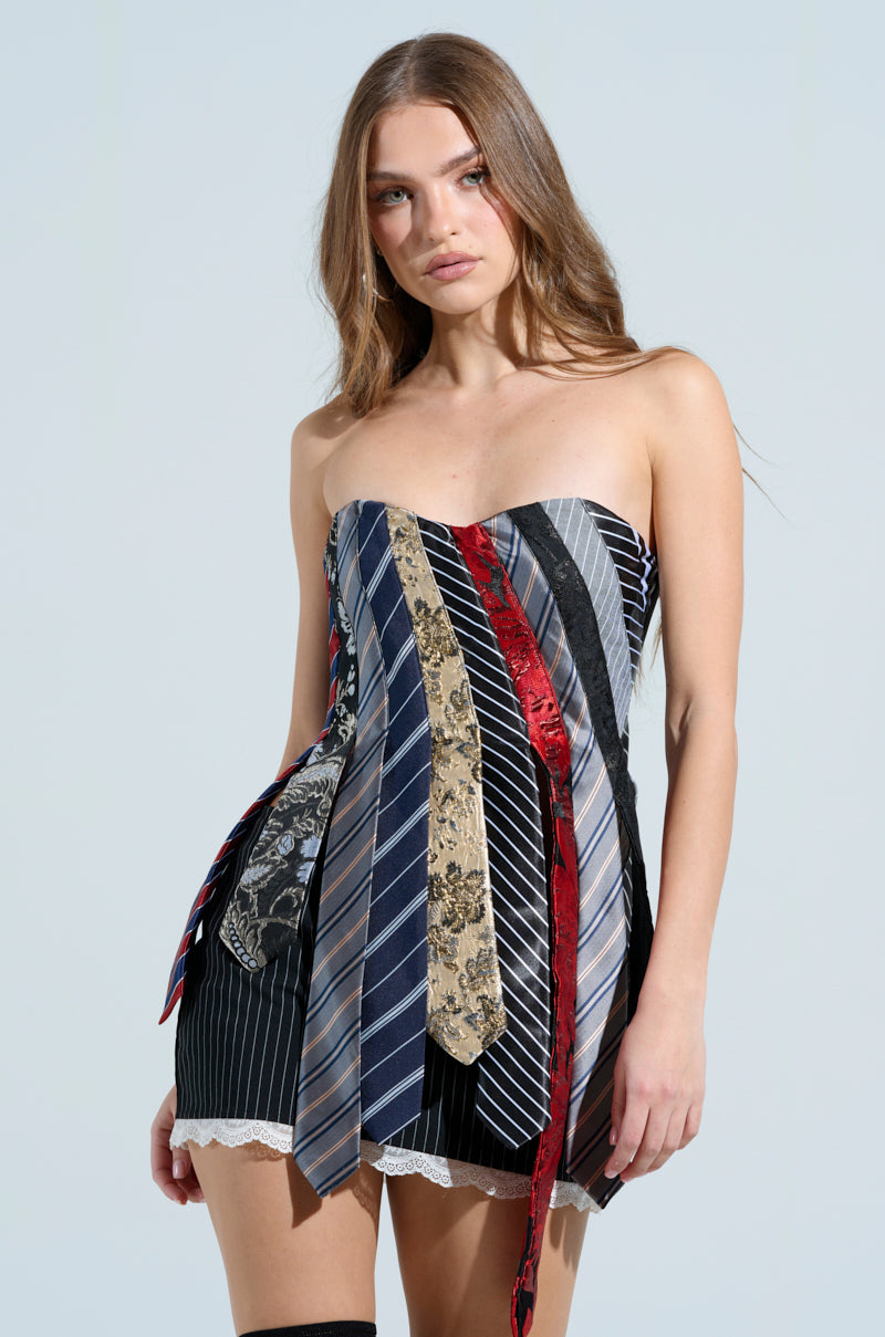 A woman with long hair poses in the STRICTLY BUSINESS TIE DETAIL CORSET TOP, featuring neckties with unique patterns and colors, including floral designs. The top showcases a strapless neckline and an asymmetrical style, blending elegance and business casual flair against a plain background.