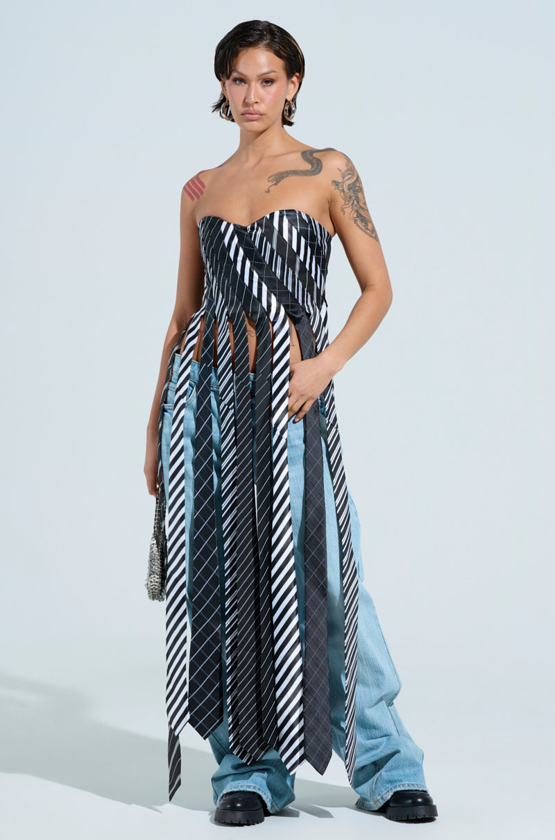 In a light blue setting, a short-haired individual poses confidently wearing the ALL WORK AND NO PLAY TIE DETAIL CORSET TOP with a strapless sweetheart neckline and black-and-white stripes. They're paired with loose blue jeans and black shoes, showcasing tattoos on their arm and shoulder.