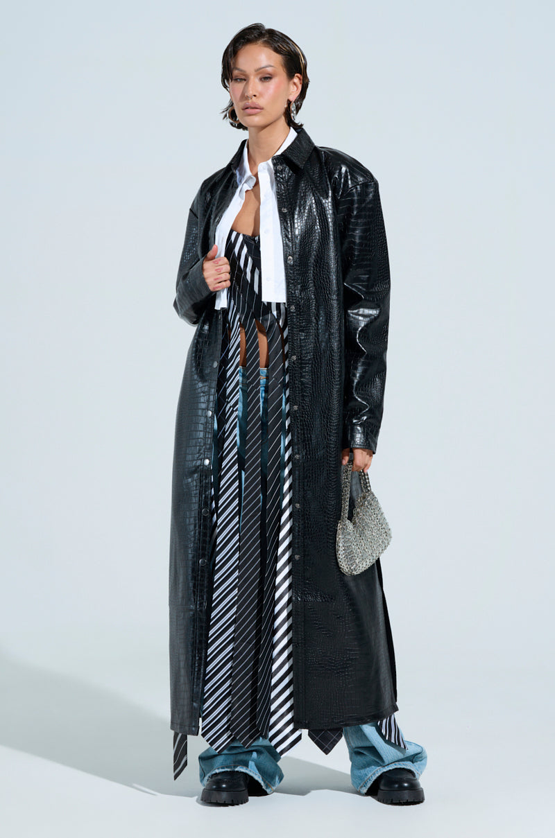 A person poses in a black faux leather trench coat over the "All Work And No Play" tie detail corset top and denim pants. They hold a sparkling handbag and wear chunky black boots, creating a stylish, modern look against a light blue backdrop.