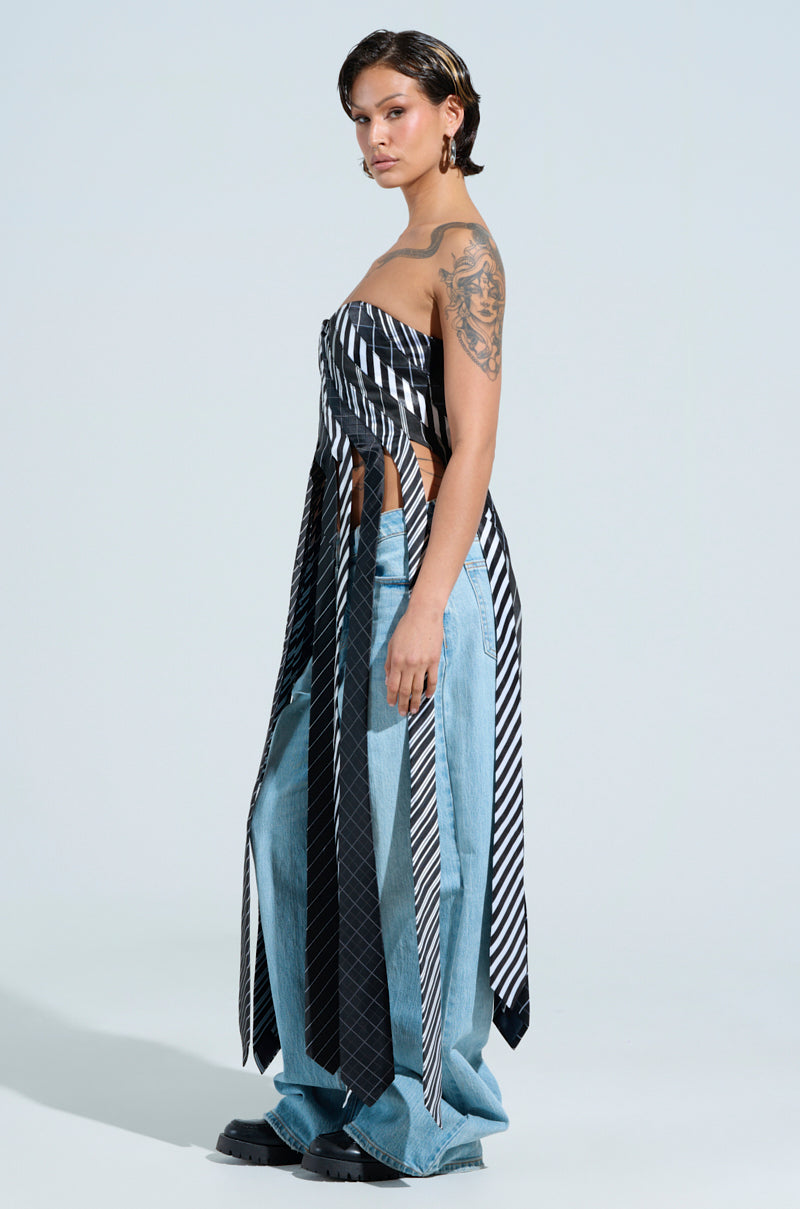A person with short hair stands confidently in a side profile, wearing the ALL WORK AND NO PLAY TIE DETAIL CORSET TOP with wide-leg jeans and chunky shoes, blending business casual elements. A large tattoo graces their left arm, and they sport hoop earrings against a plain light-colored background.