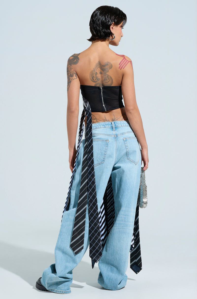 A person with short dark hair stands with their back to the camera, wearing the ALL WORK AND NO PLAY TIE DETAIL CORSET TOP and light blue jeans with ties hanging down. Holding a small beaded bag, they reveal tattoos on their upper back and arm.